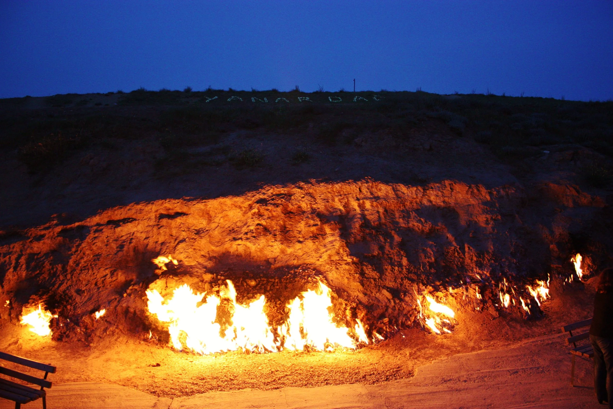 A hillside on fire during dusk with flames burning along the base, illuminated by an orange glow, and the sky fading to dark blue, with a partially visible bench on the right and the word 'LANTERN' spelled out in large letters on the hilltop.