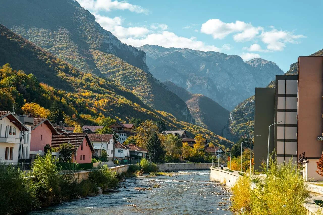 A small town with colorful houses next to a river, surrounded by mountains with green and yellow foliage during fall.