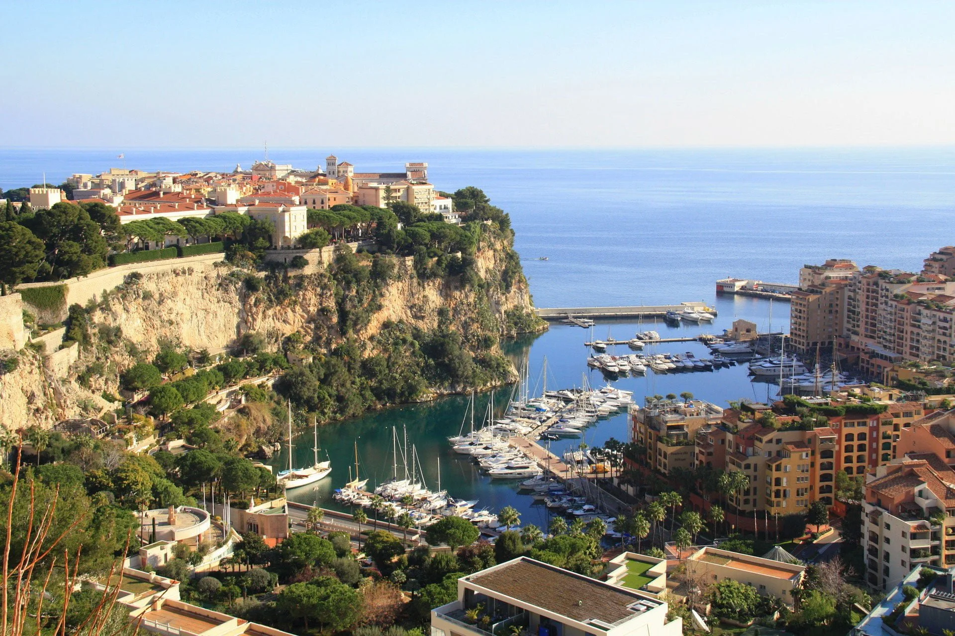 A scenic view of a coastal city with a marina full of sailboats, surrounded by colorful buildings, lush greenery, and a cliff overlooking the ocean.