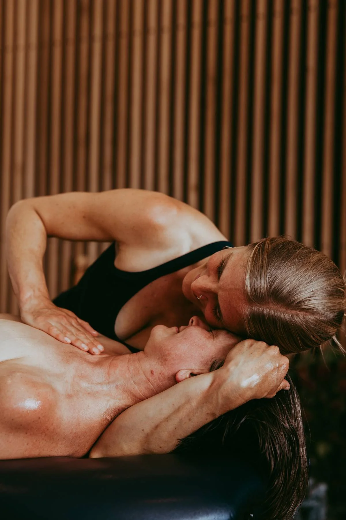 A woman is giving a passionate kiss to a man lying down, with her hands touching his face and neck, against a wooden backdrop. 滋養、神聖空間、連結