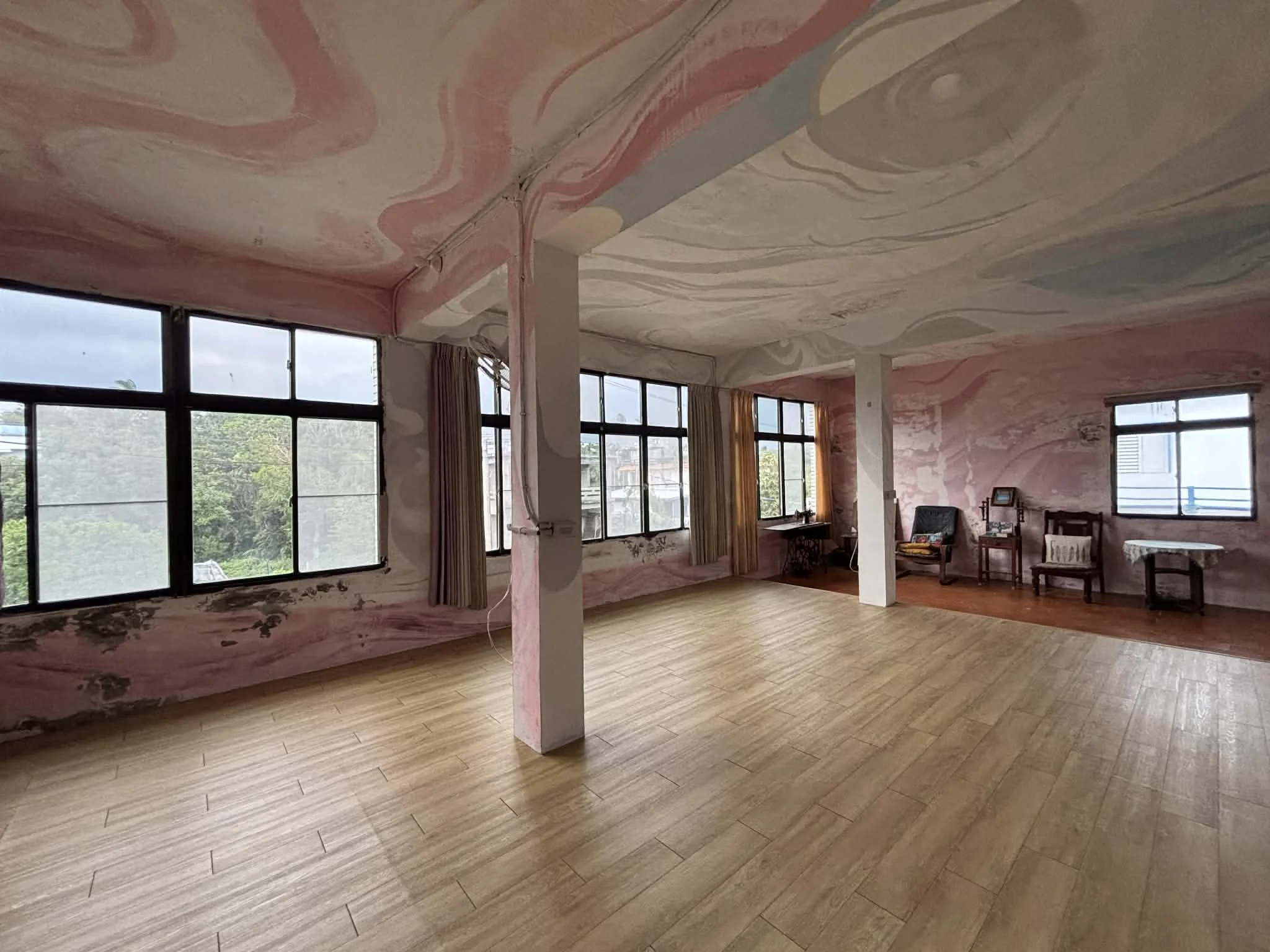Empty room with large windows, wood flooring, pink marbled walls and ceiling, and vintage chairs along the wall.