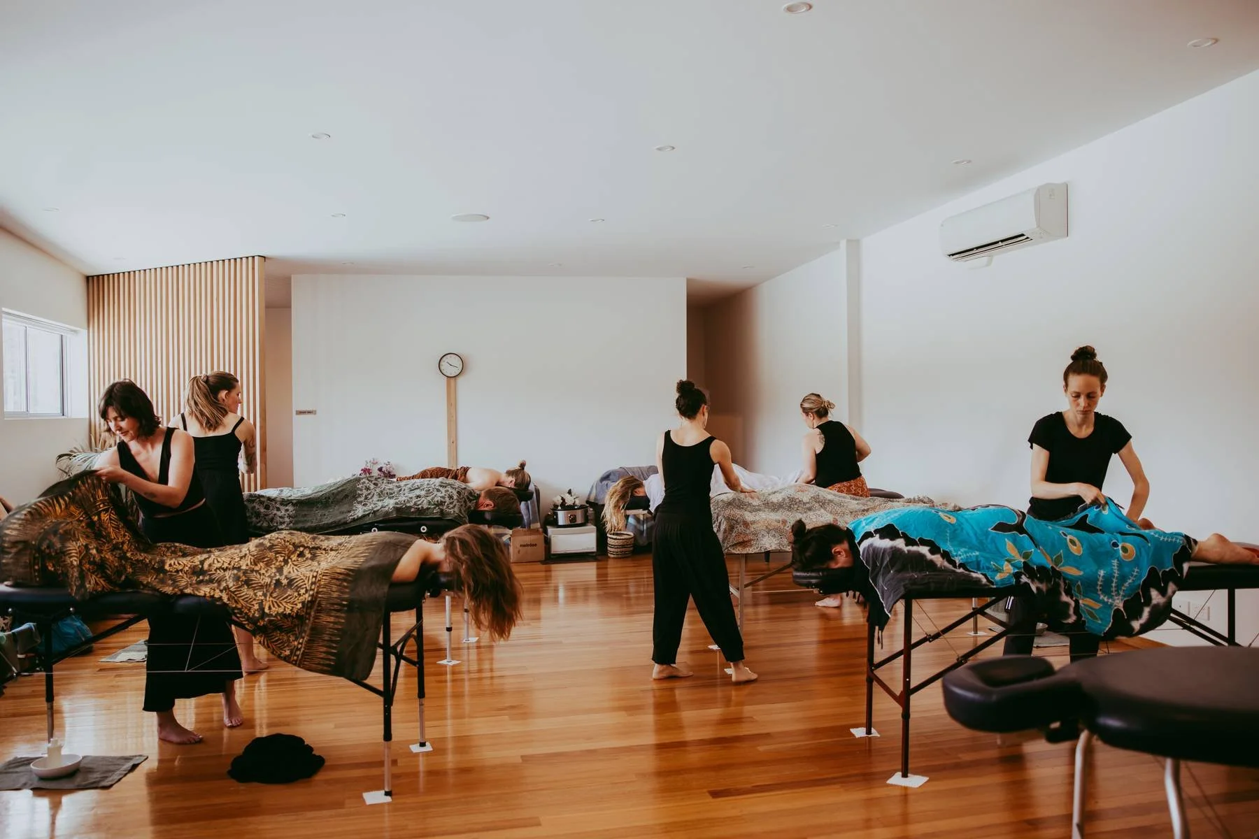 People receiving massages in a spacious, well-lit room with wooden floors, some lying on massage tables and others standing, with a clock on the wall and an air conditioner mounted above.