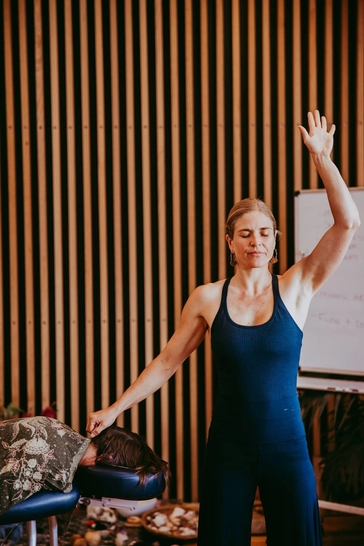Woman with closed eyes, dressed in fitness attire, practicing a yoga pose in a studio with a woman resting face down on a massage table, background includes wooden wall panels and a whiteboard. 祈禱、夏威夷、連結、療癒、神聖、按摩