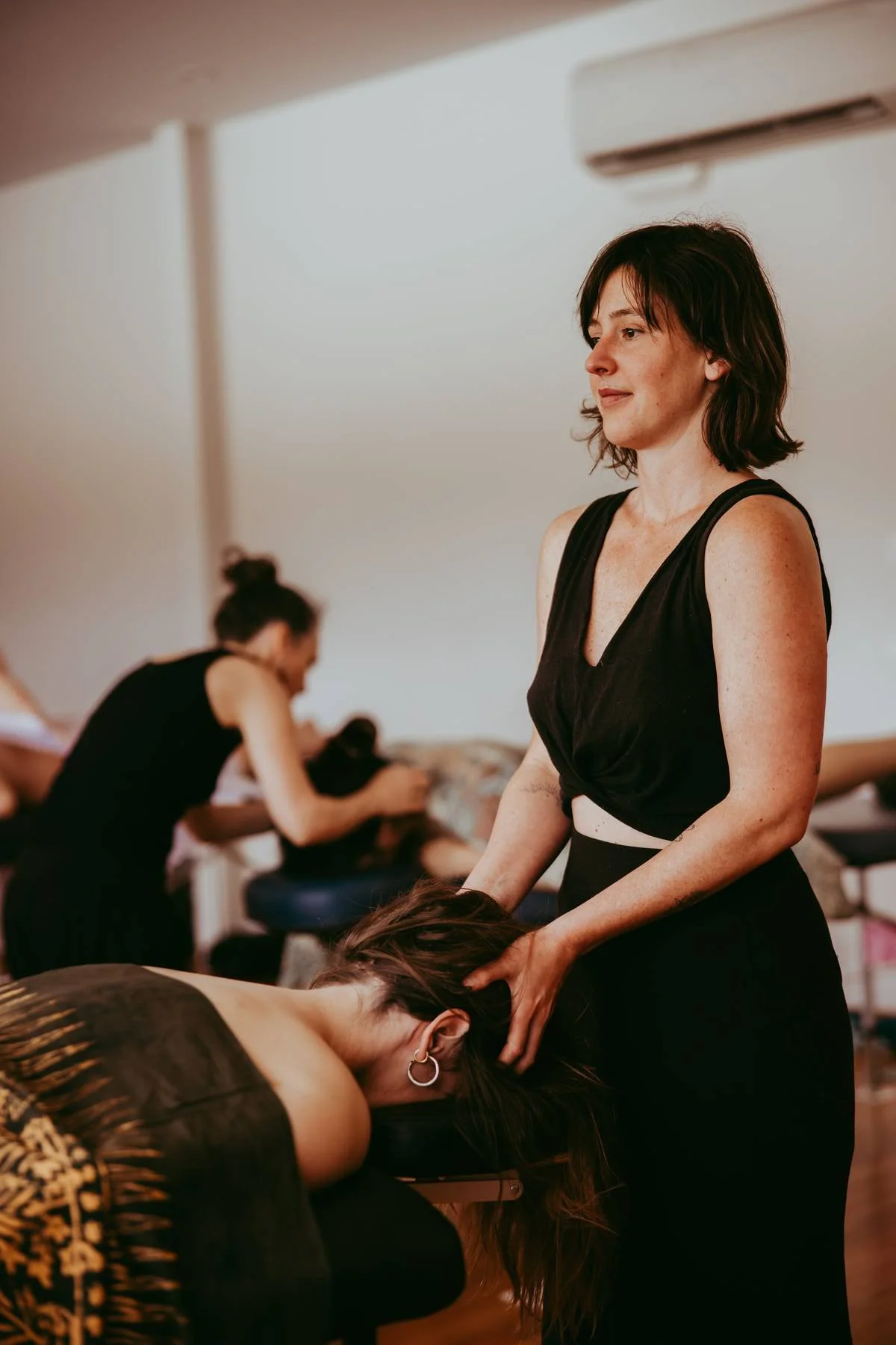 A woman giving a head massage to another woman lying face down on a massage table, with two other women in the background preparing for their massages.