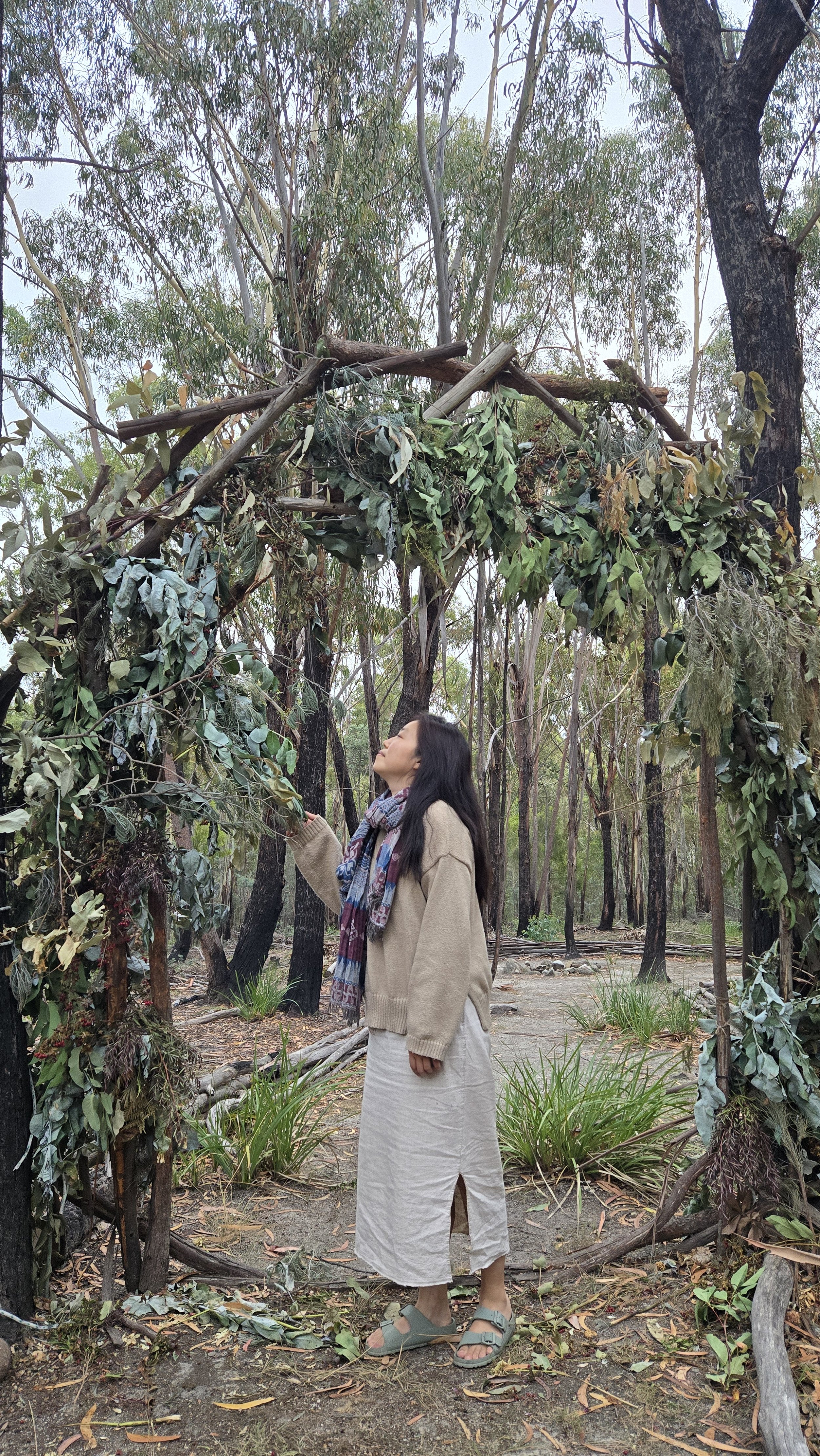 A woman standing outdoors in a wooded area with a natural archway made of sticks and greenery above her. She is wearing a beige sweater, a long skirt, sandals, and a colorful scarf. @oneheart.bodywork