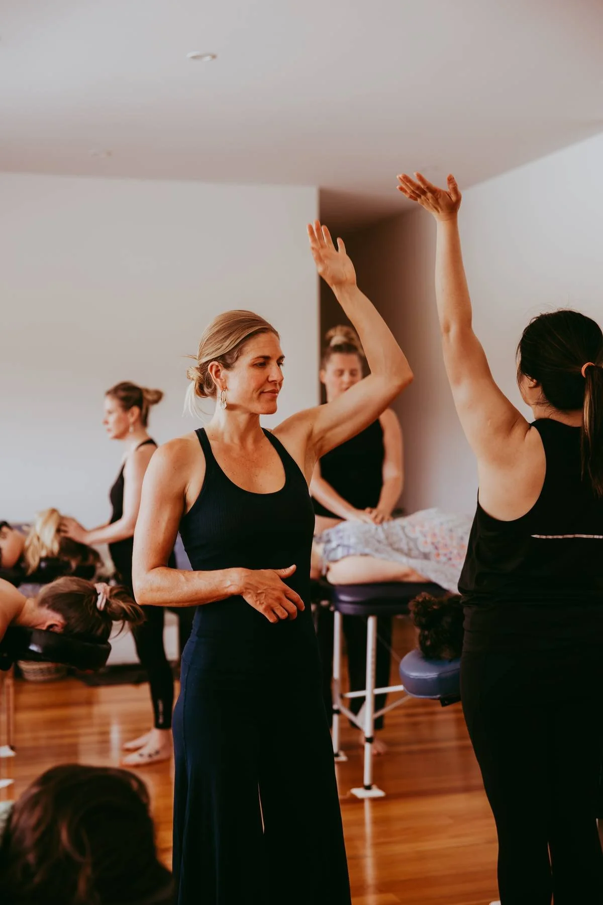 Women participating in a yoga or wellness class, with two women exchanging high-fives in the foreground and others stretching or resting in the background.