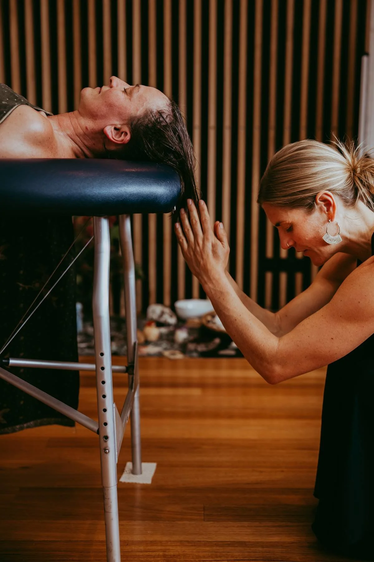 A woman practicing yoga on a massage table with another woman in a yoga studio with wood panel walls.