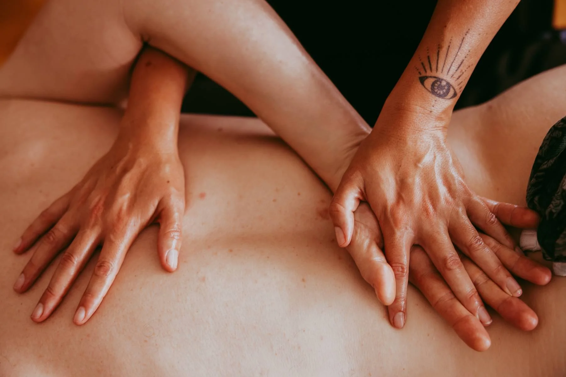 Close-up of a massage therapist's hands applying a massage to a person's back, with a tattoo of an eye on one finger.