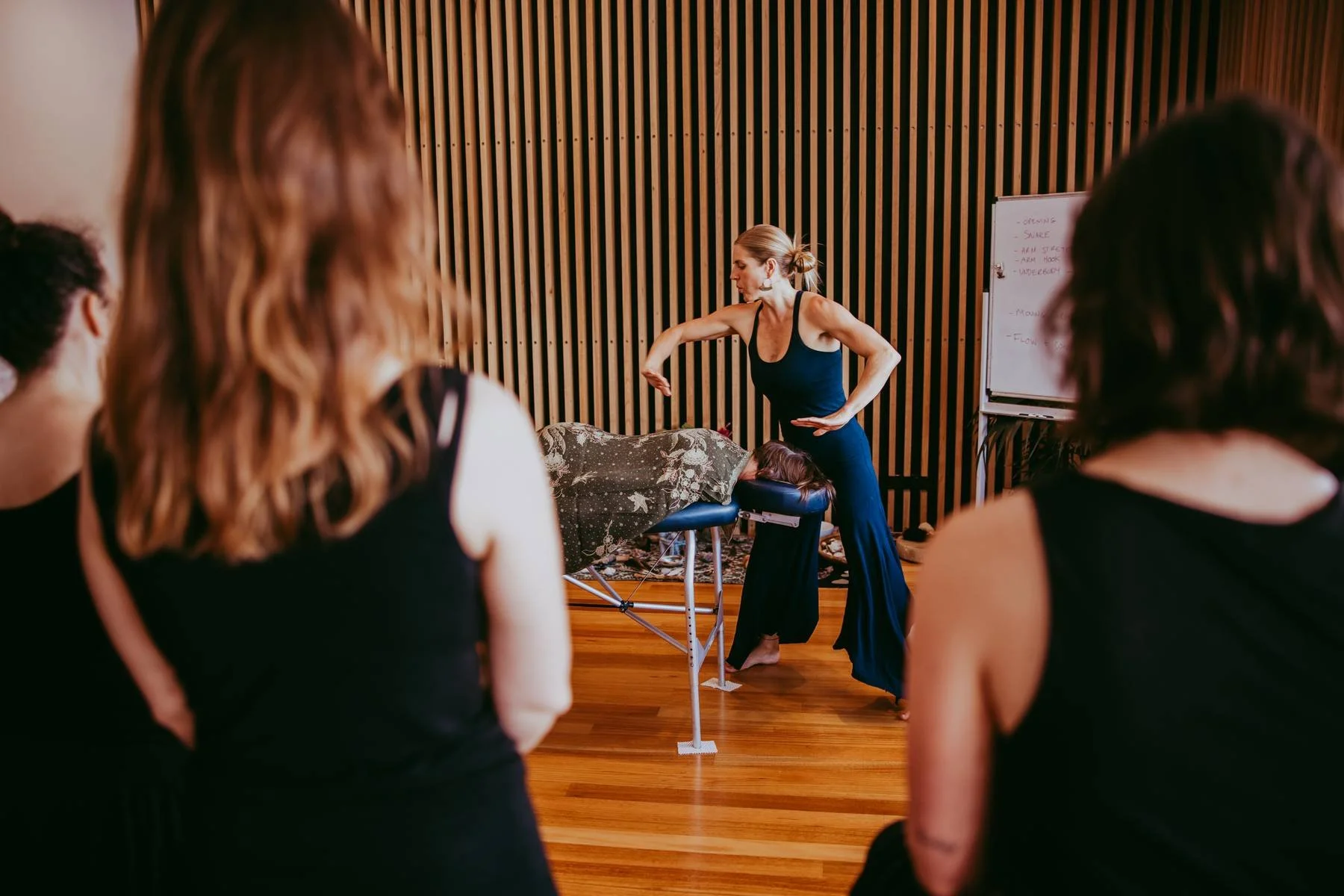 A woman giving a massage demonstration to a small group in a room with wooden walls and floor. Four women are observing, with their backs to the camera, focusing on the instructor.