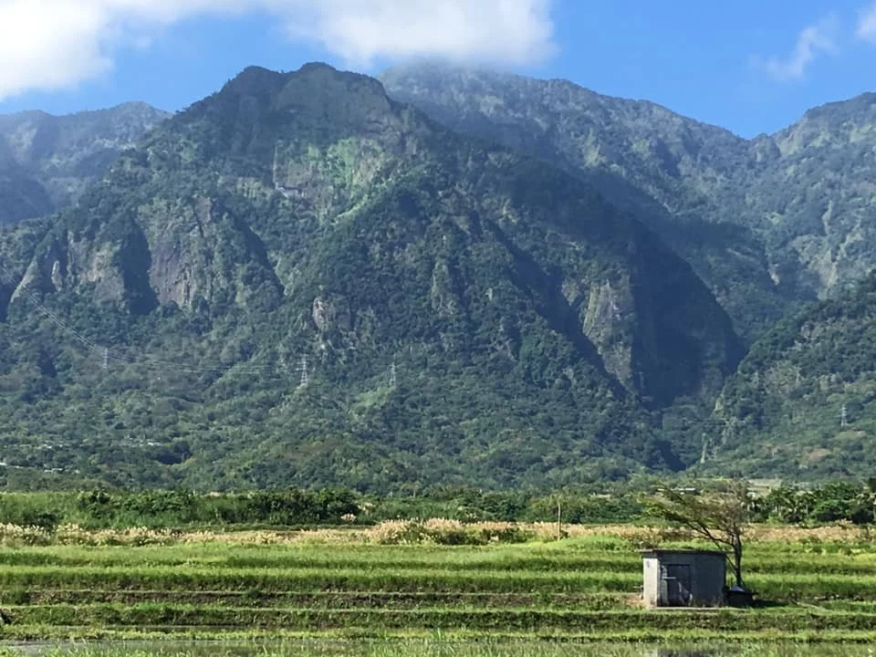 A mountain with green vegetation, a small building, and flat farmland in the foreground.