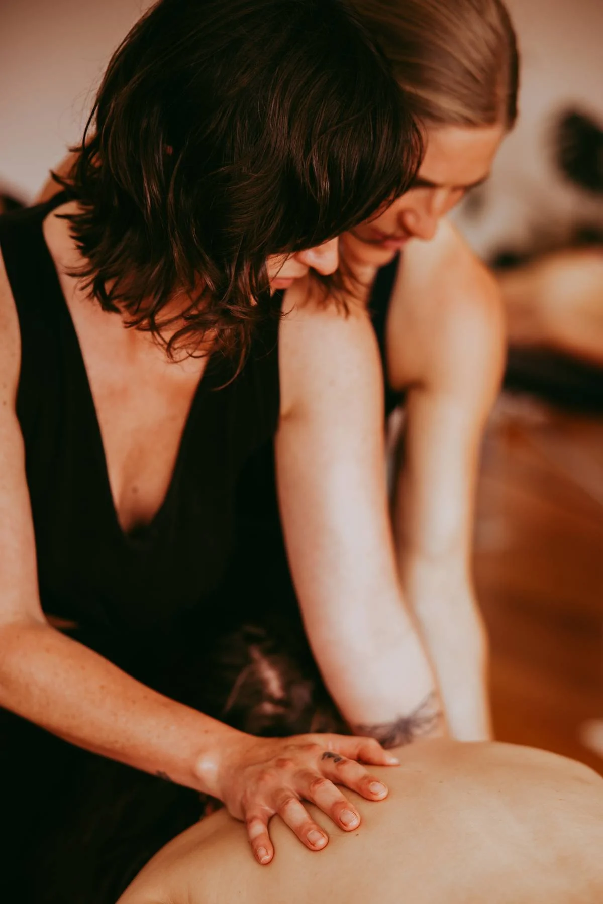 Two women giving a massage, one with dark curly hair and the other with light brown hair, in a warm, softly lit room.
