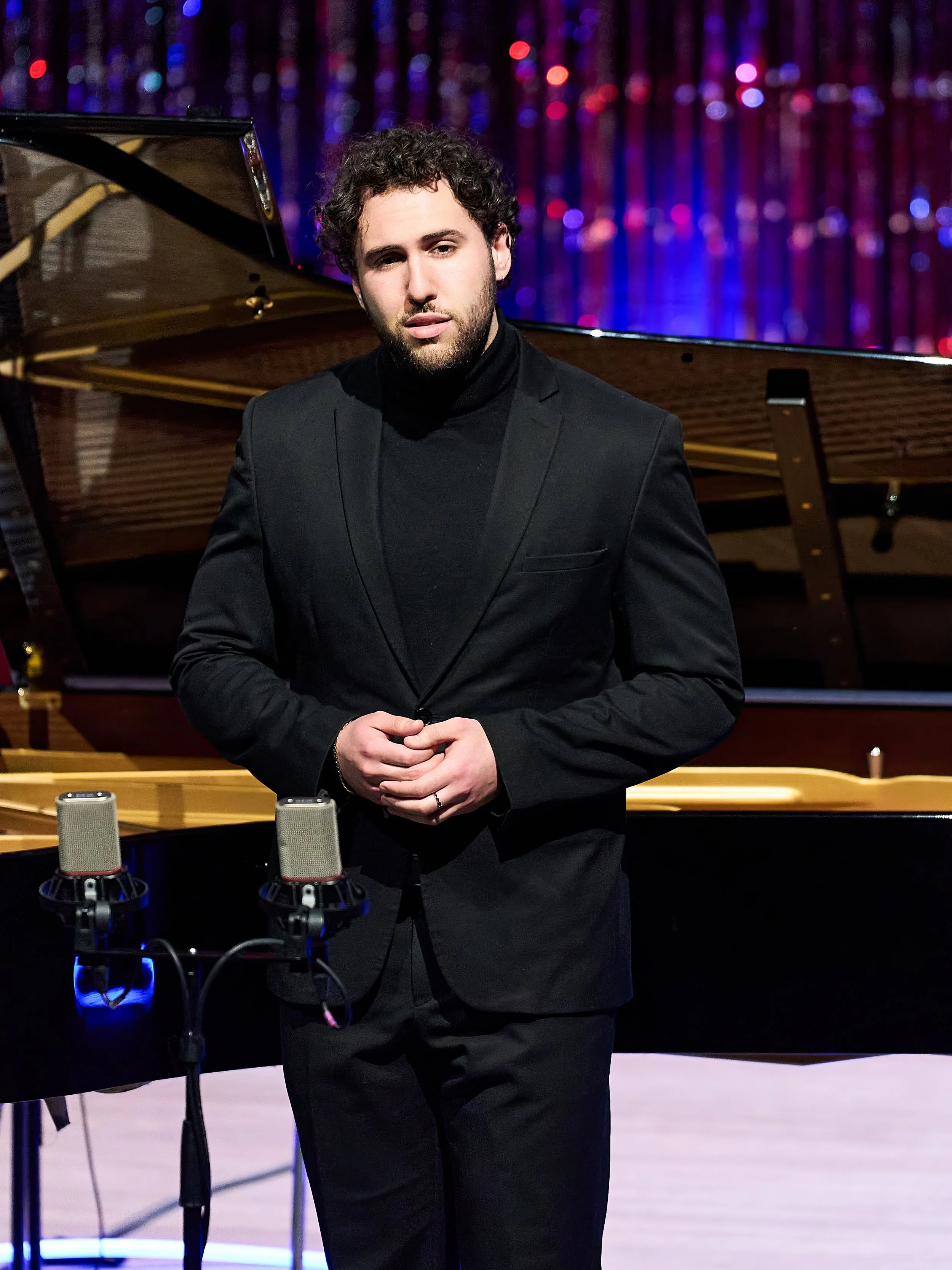 A man in a black suit and turtleneck standing in front of a grand piano with a background of colorful, shimmering curtains.