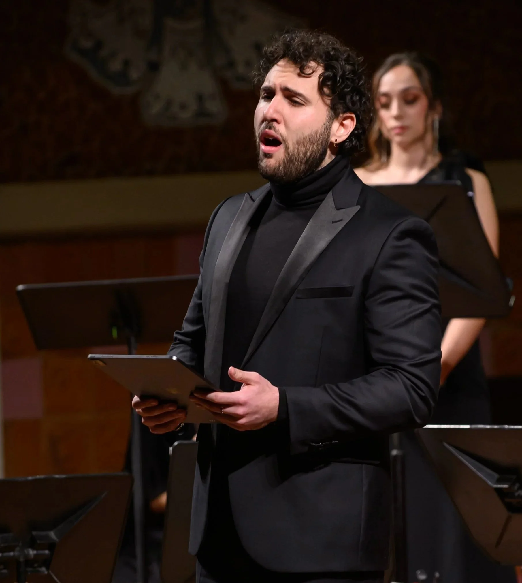 Man singing passionately during a performance, holding a tablet, with a woman playing the piano in the background.
