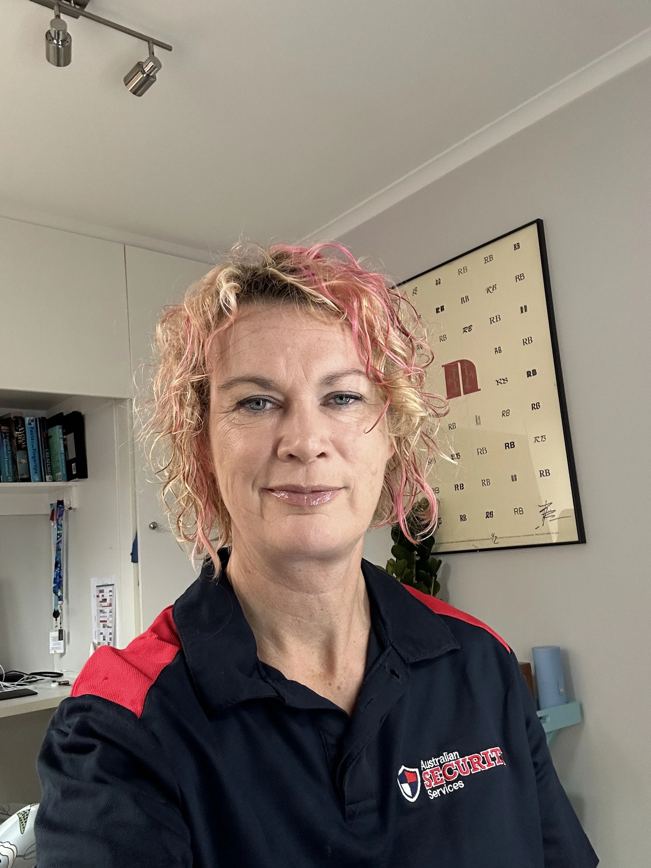 A woman with curly blonde and pink hair taking a selfie indoors, wearing a black polo shirt with red accents and the logo "Australian Security Services" on it.