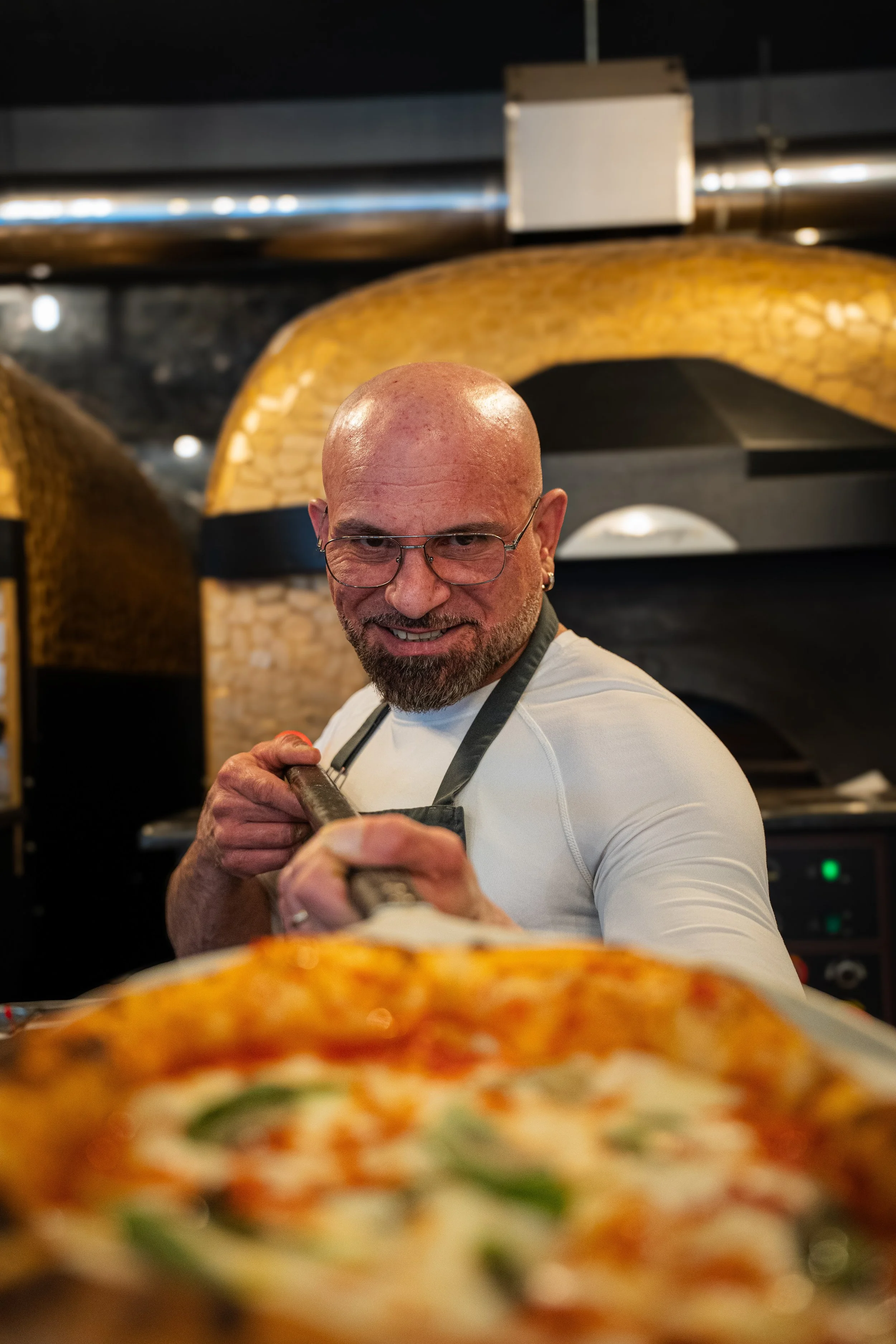 Mann - Francesco Ialazzo, mit Glatze, Brille und Bart, der eine Pizza schneidet, vor einer Pizza in einem Restaurant mit Backöfen im Hintergrund.