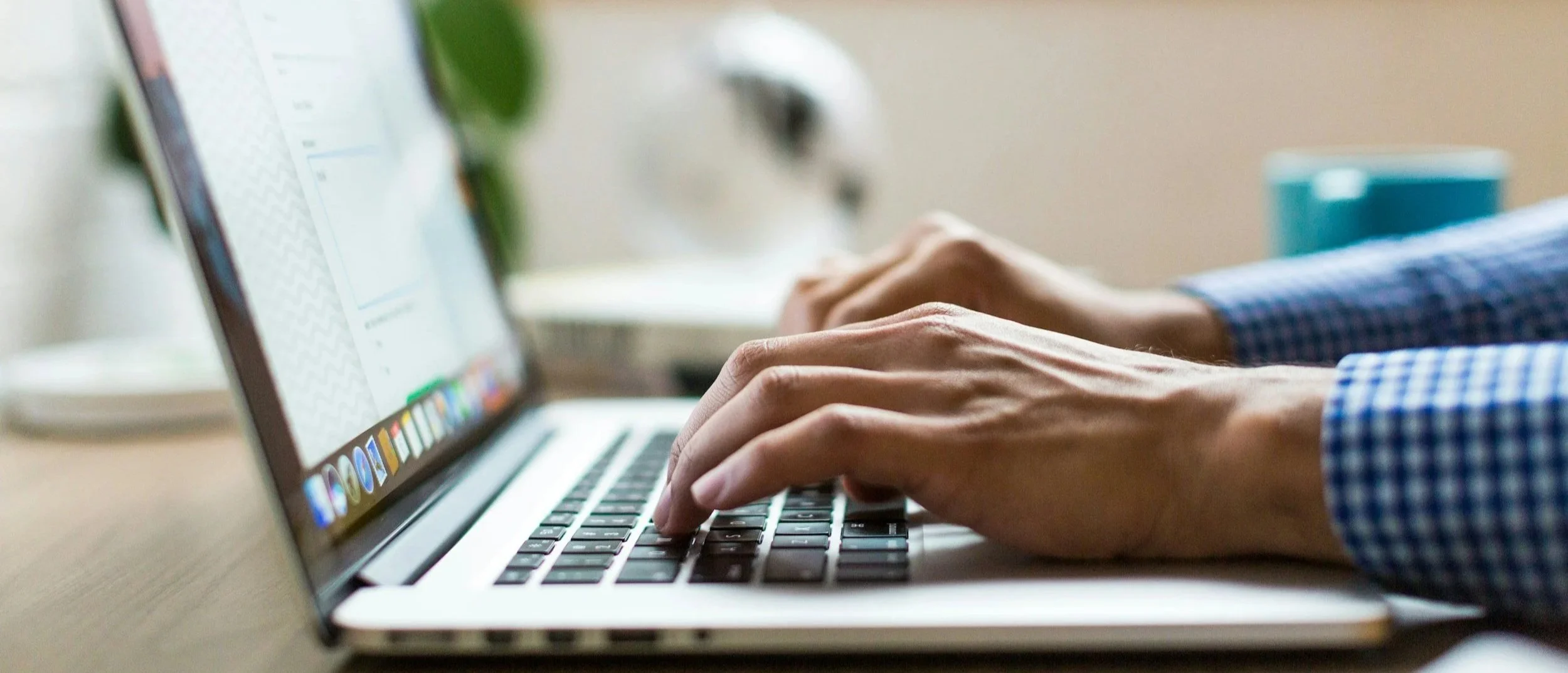 Close-up of someone typing on a laptop keyboard with a blurred dog and a coffee mug in the background.