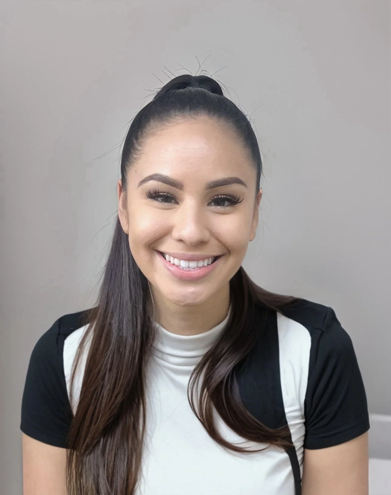 A young woman with long dark hair in a high ponytail, smiling, wearing a black and white short-sleeved shirt, sitting against a plain light gray background.