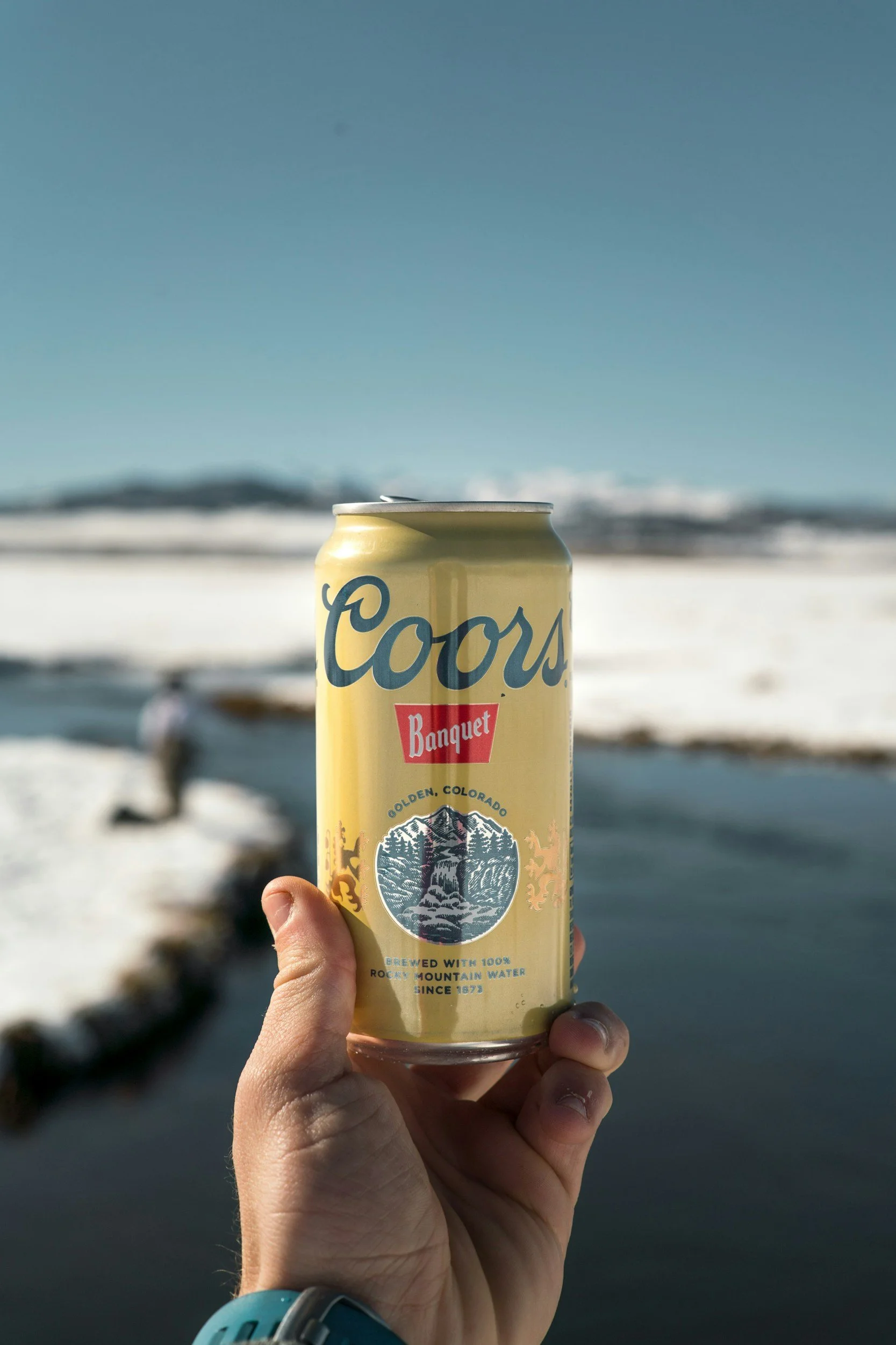 Eine Hand hält eine Dose Coors Banquet Bier vor einer Winterlandschaft mit Schnee, einem Fluss und Bergen im Hintergrund.