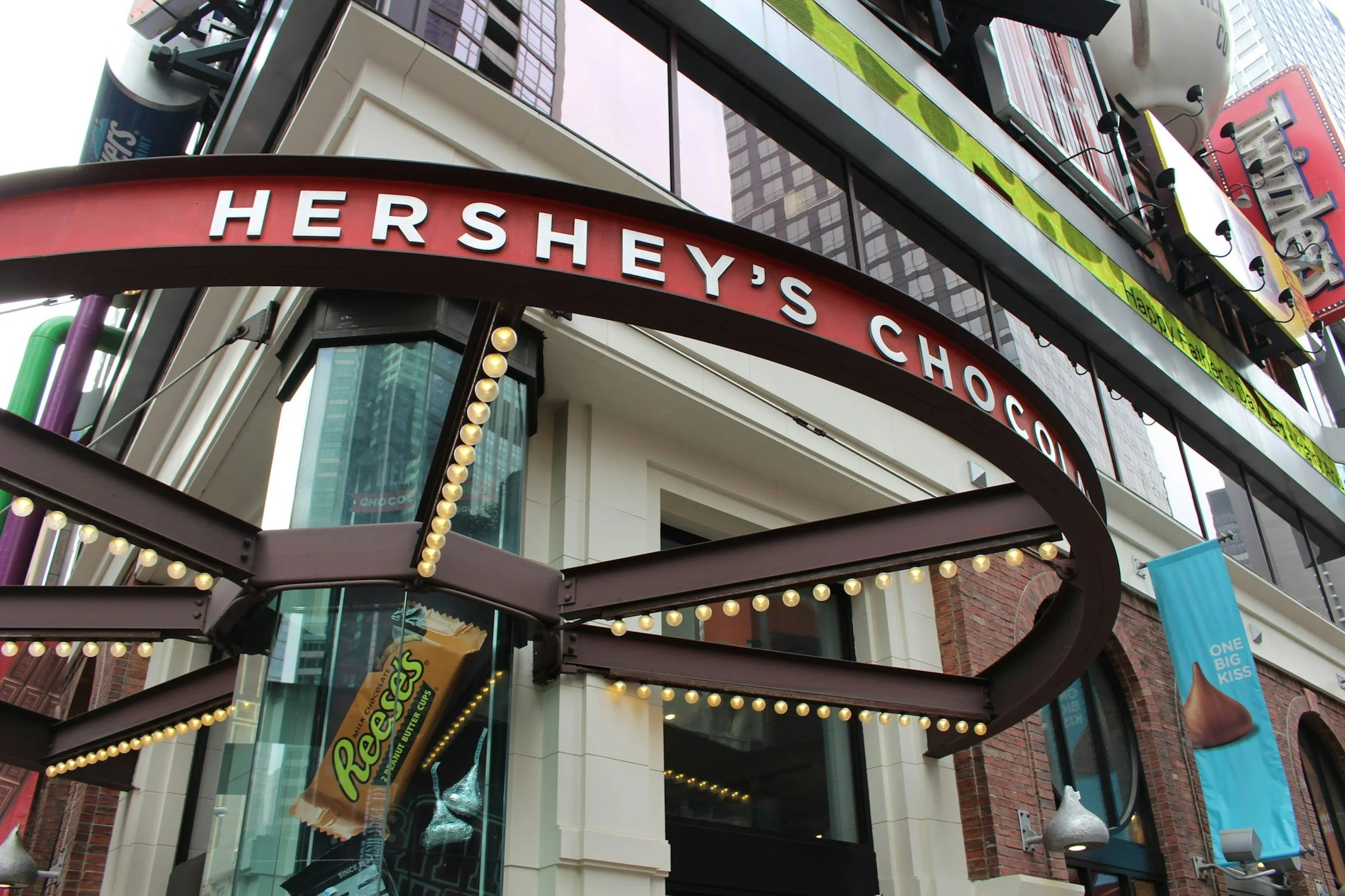 Außenansicht eines Hershey's Chocolate-Geschäfts mit leuchtendem Schild und Glühbirnen, Hochhäuser im Hintergrund.