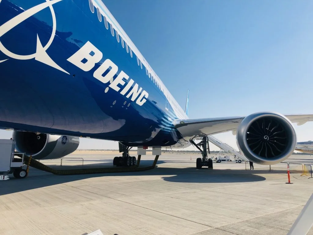 Large Boeing airplane on airport tarmac with blue fuselage and white Boeing logo, jet engine visible on the right, clear sky in the background.