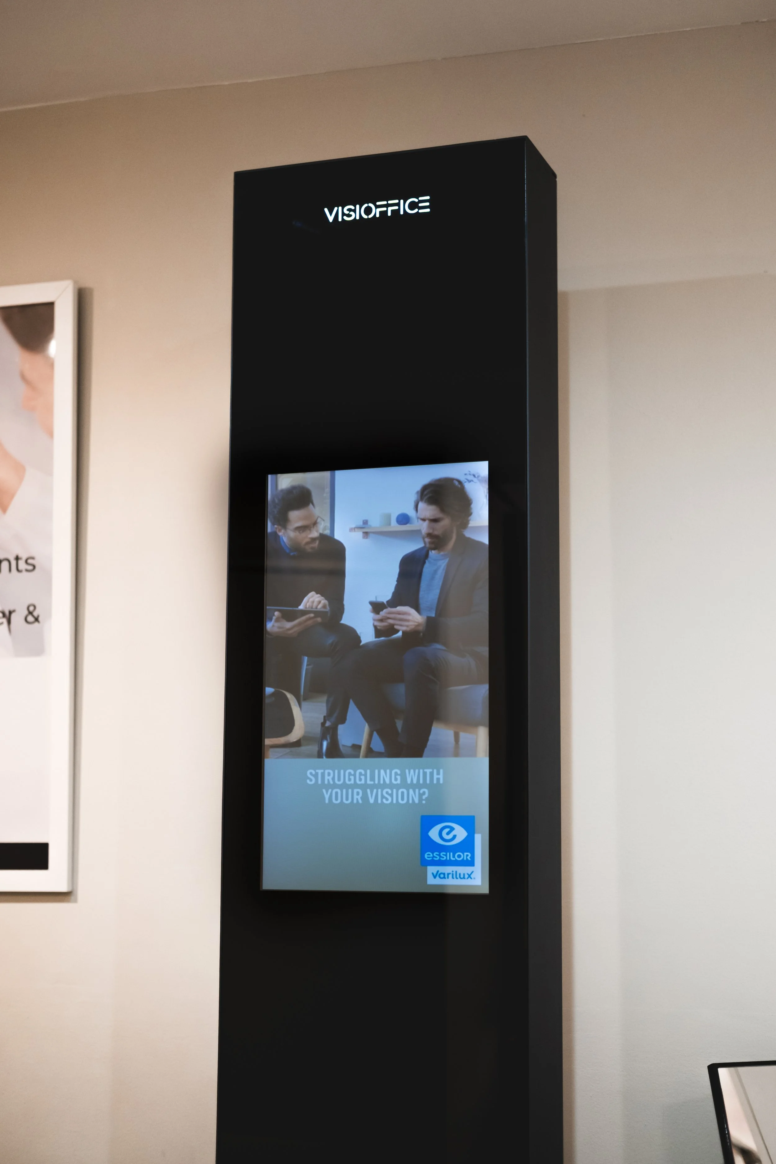 Digital display on a black kiosk featuring two men in suits looking at a phone with the text 'Struggling with your vision?' and logos for Essilor and Varilux.