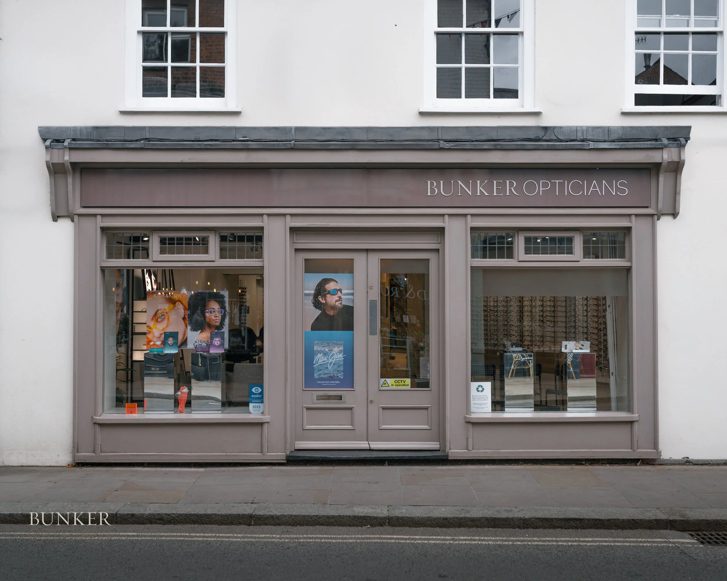 Exterior of an optical store named BUNKER OPTICIANS with large display windows and a central door, showing promotional posters for glasses and eyewear inside.