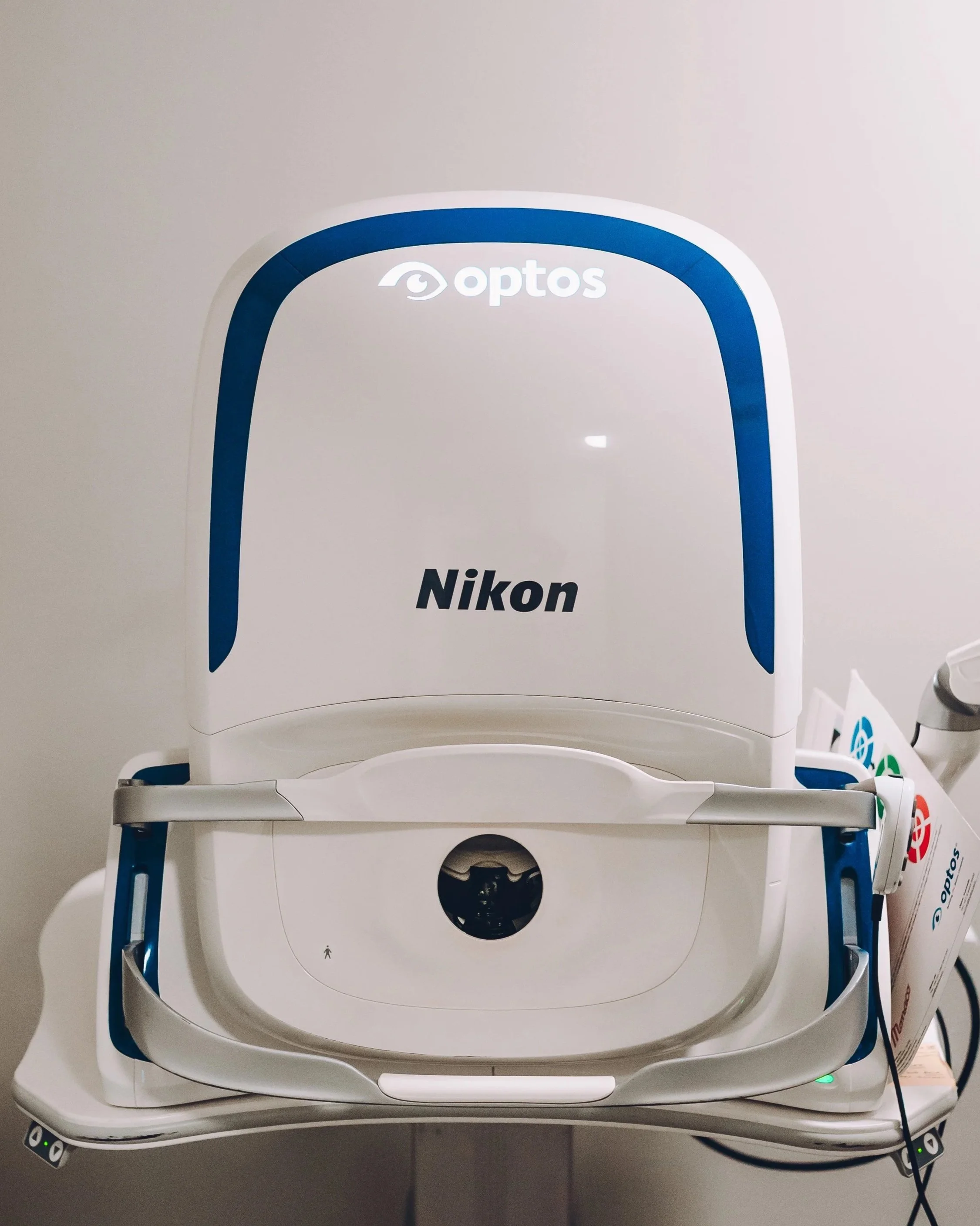 An eye examination machine labeled 'Optos' and 'Nikon' in a clinical setting.