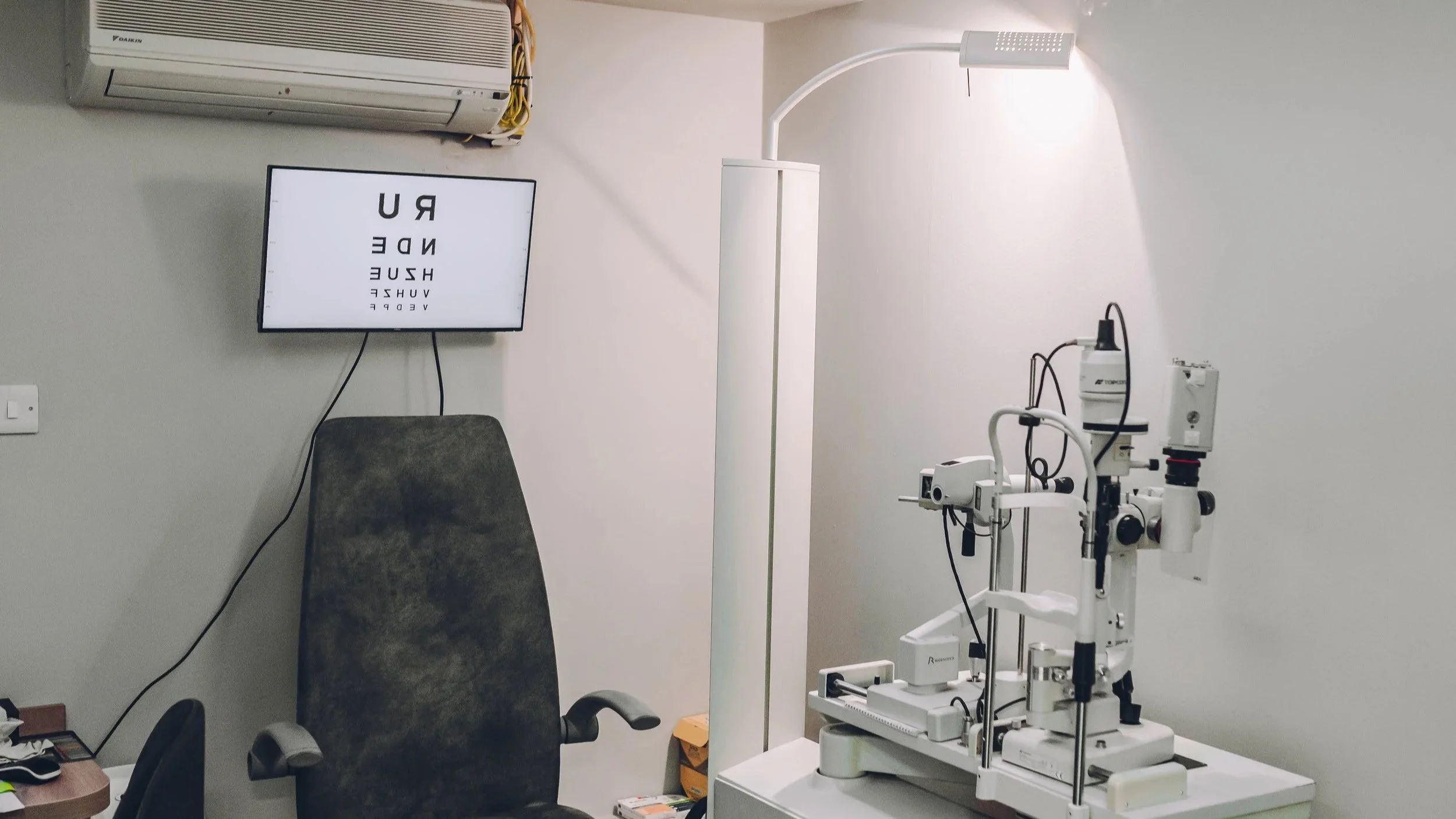 Medical examination room with an eye chart on a mounted screen, an operating microscope on a table, a chair, and an air conditioning unit on the wall.