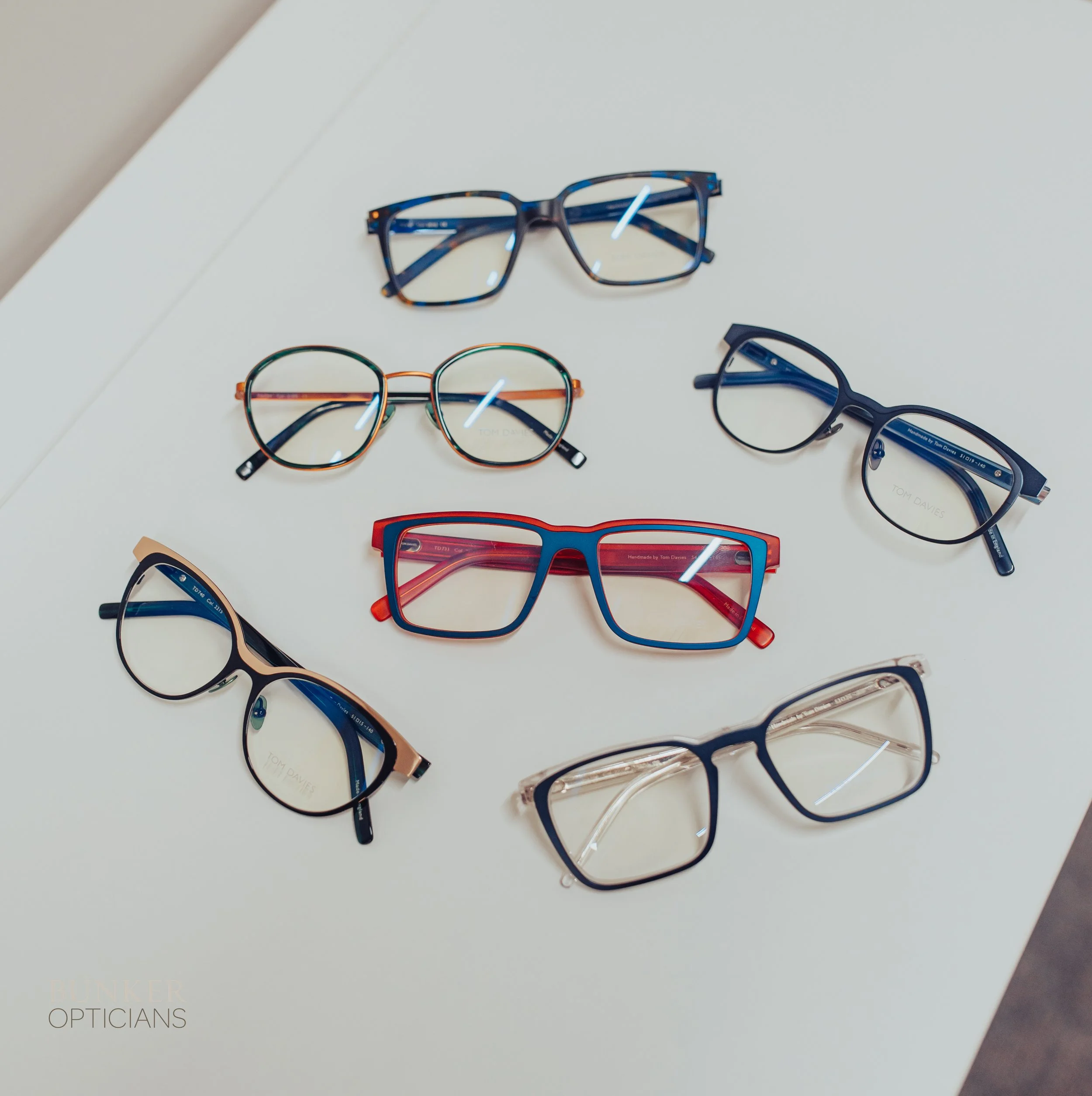 Six pairs of eyeglasses arranged on a white surface with the words "TINKER OPTICIANS" in the bottom left corner.
