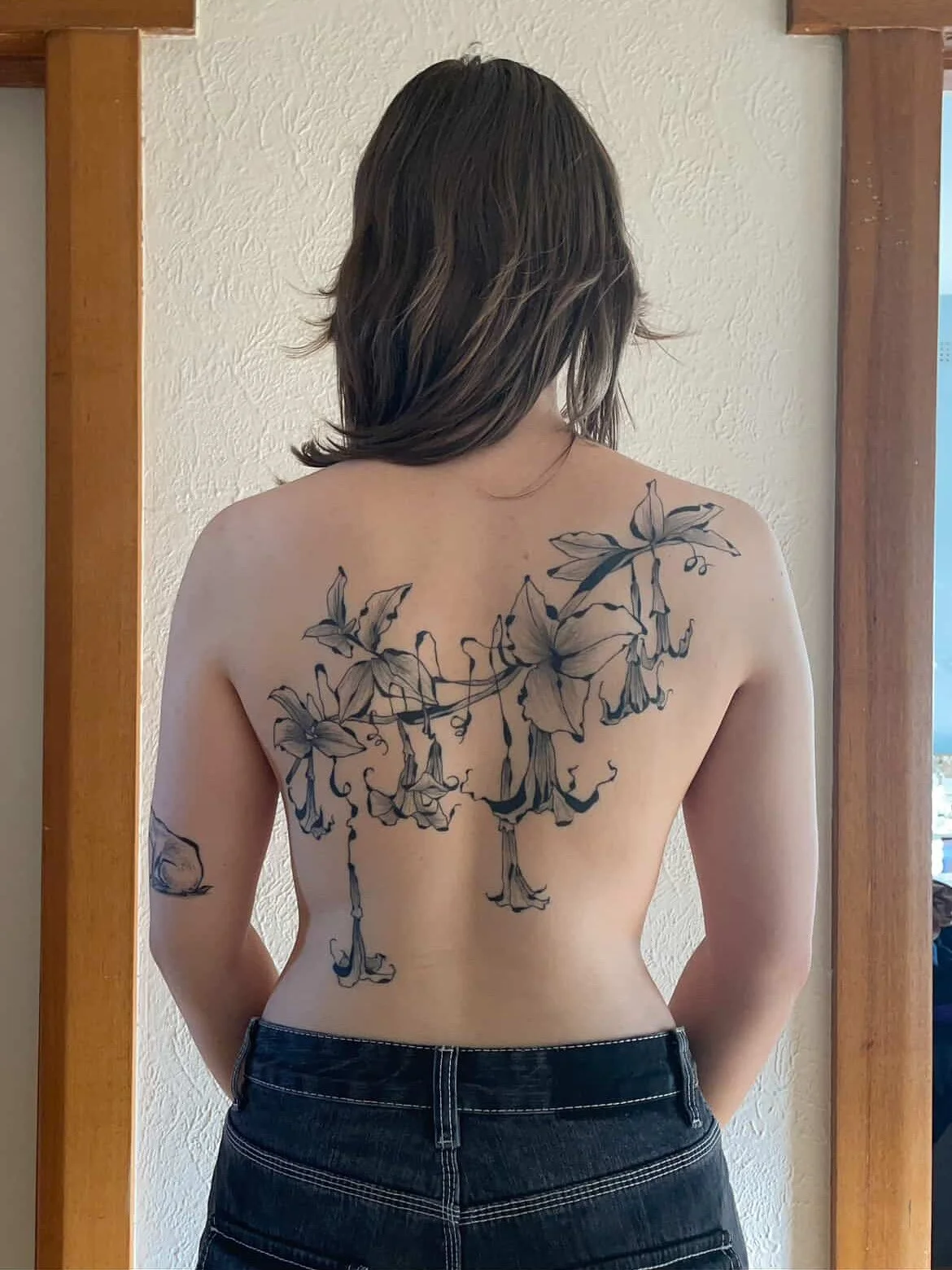 Person with shoulder-length hair showing a large floral tattoo on their back, wearing dark jeans, standing indoors in front of a mirror.
