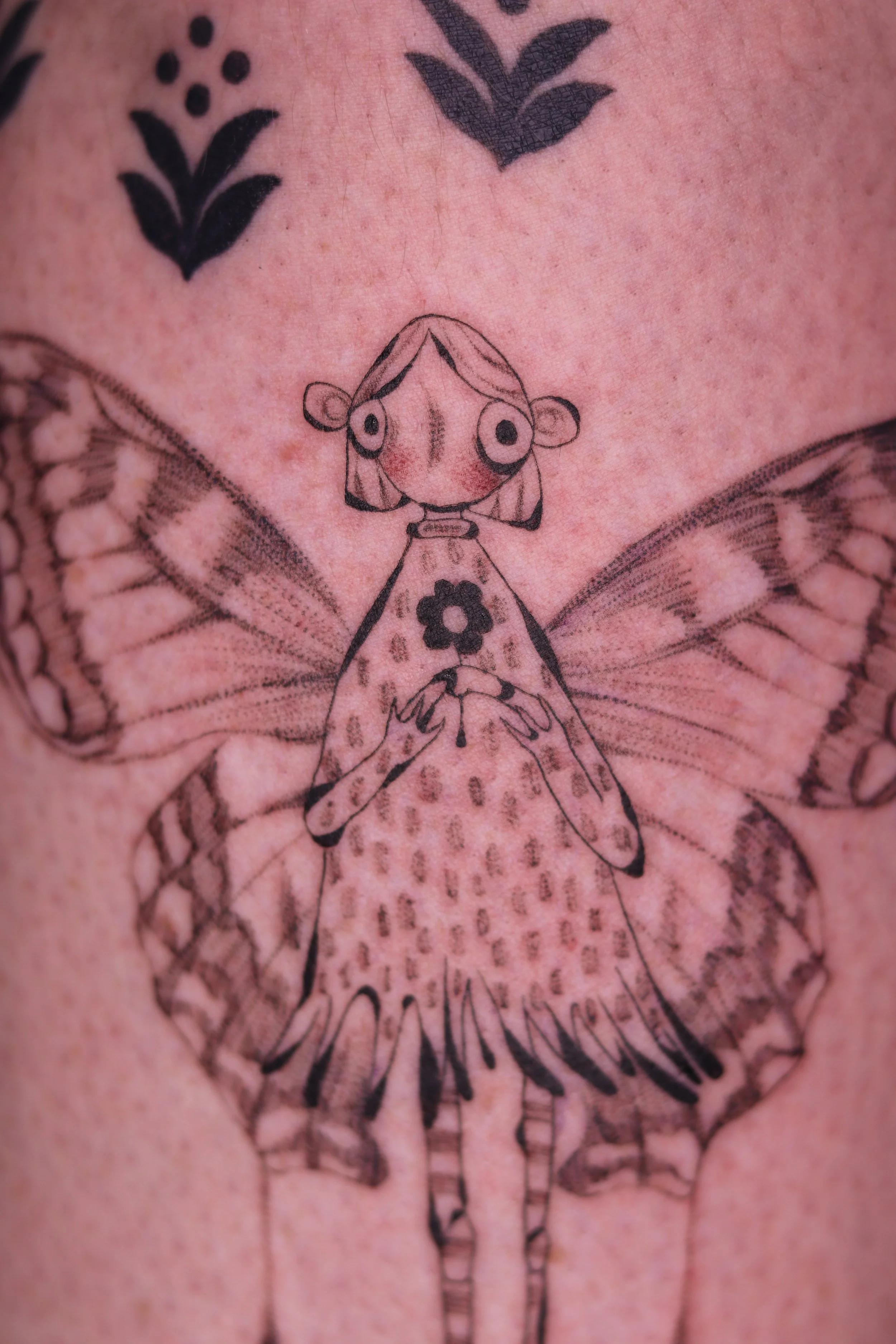 Tattoo of a cartoonish fairy with butterfly wings, big eyes, and a cute expression, on person's skin.