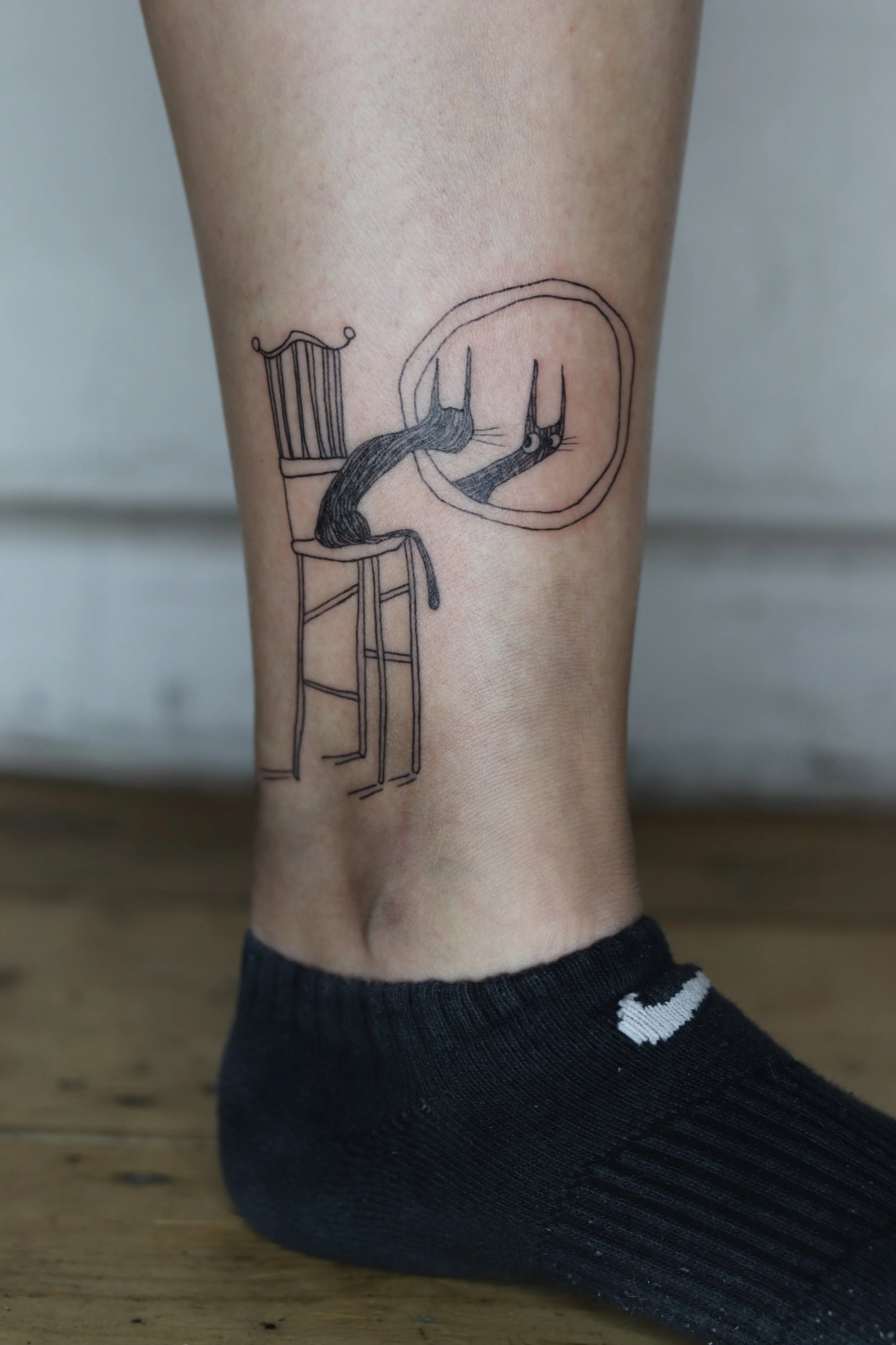 Tattoo of a cat sitting on a chair, looking into a mirror, all in minimalist black ink line art style.