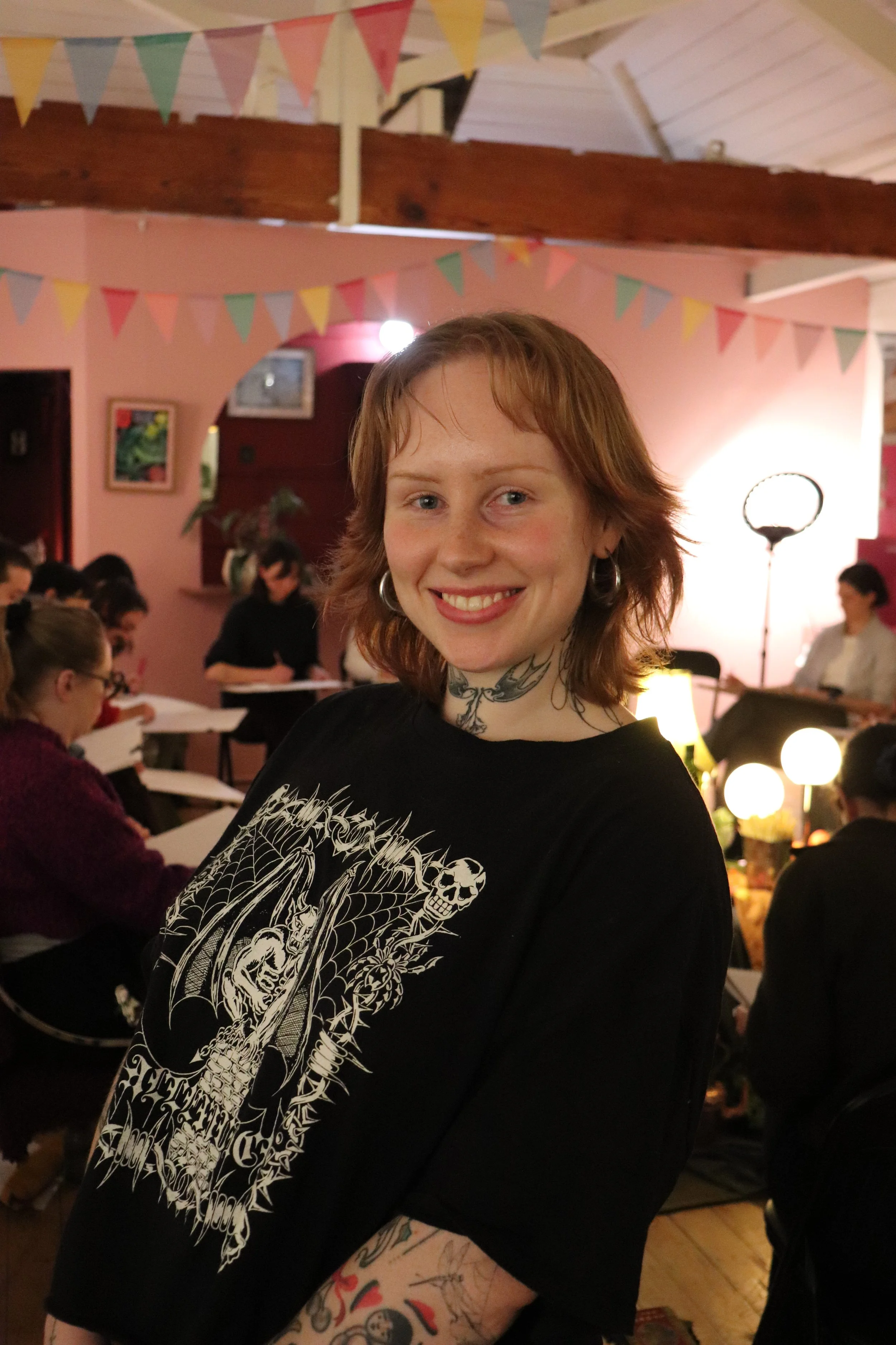A woman with short red hair, tattoos on her neck and arm, and wearing a black graphic t-shirt, smiling at a gathering in a decorated indoor space with pink walls, bunting, and seated people.