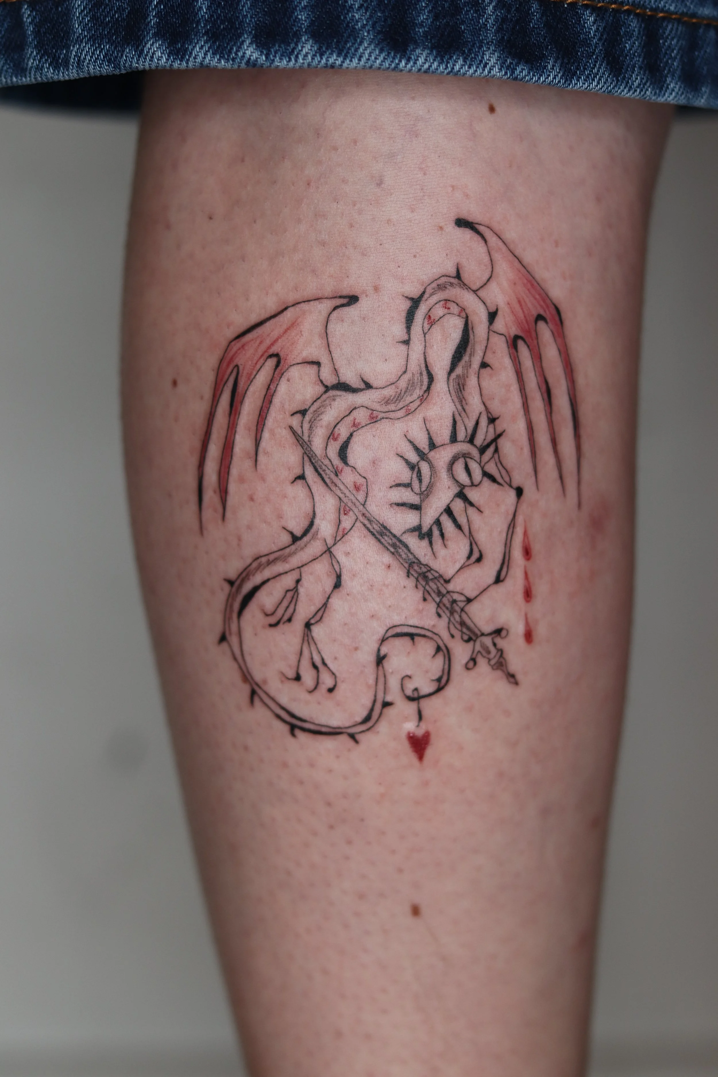 Tattoo of a dragon with wings, holding a sword, with a heart below and a sun to the side, on a person's thigh.