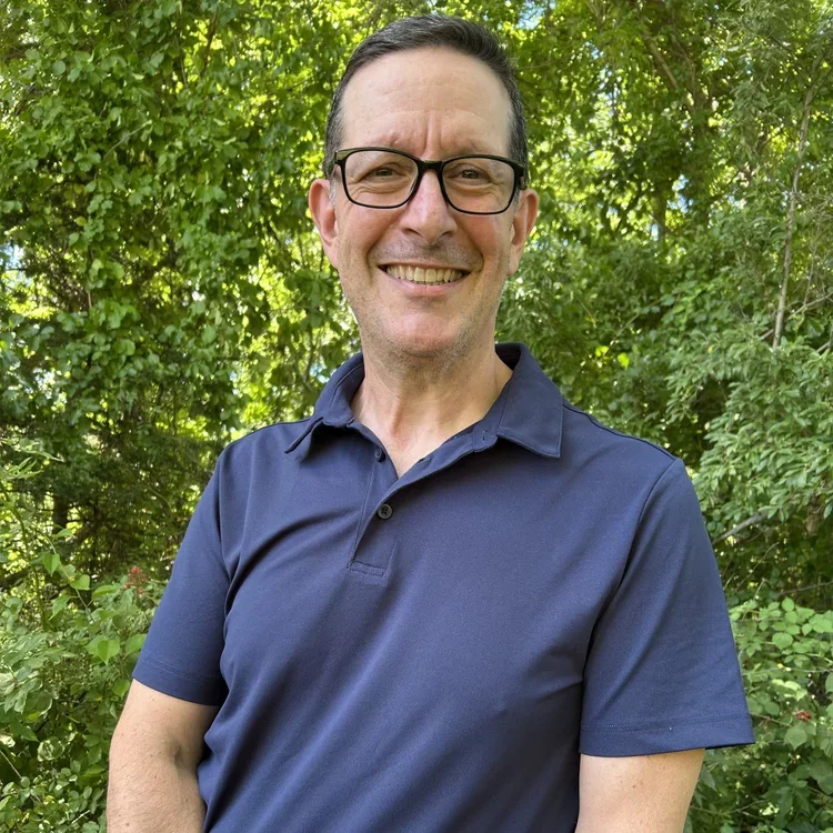 A smiling man with dark hair, glasses, wearing a navy blue polo shirt, standing outdoors in front of green foliage.