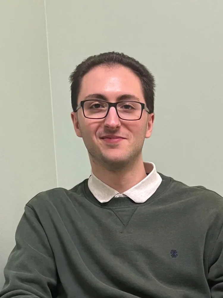 A young man with dark hair and glasses, wearing a dark green sweatshirt over a light-colored polo shirt, seated in front of a plain light green wall, smiling at the camera.