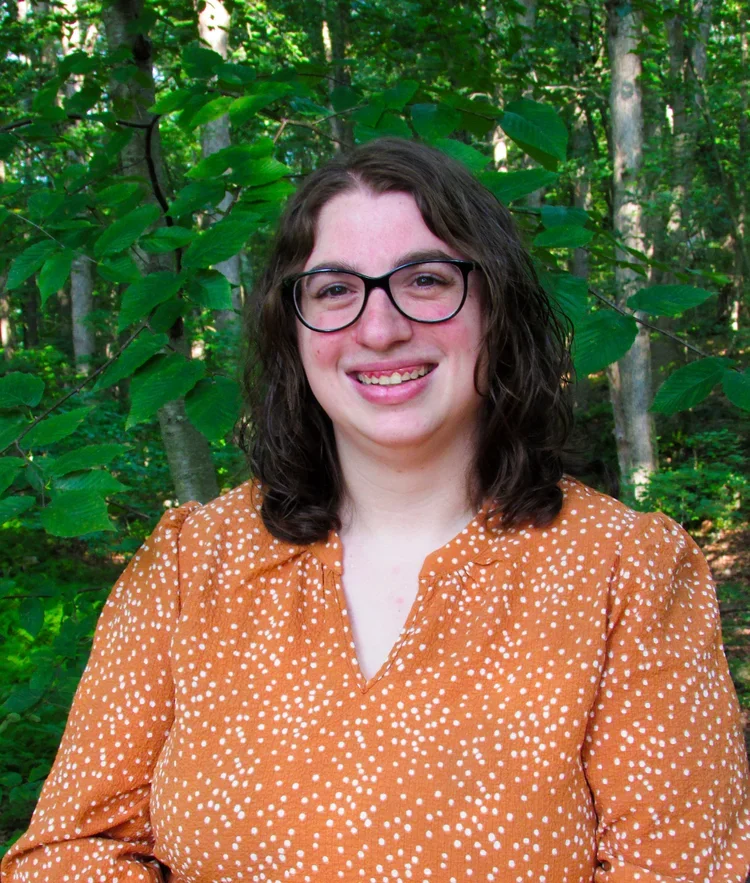 A woman with dark curly hair and glasses smiling in front of green trees in a forest.