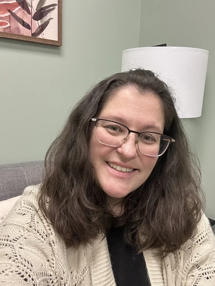 A woman with long curly brown hair, glasses, and a beige knit sweater smiling in a room with a light green wall, a white lampshade, and a framed artwork of botanical leaves in the background.