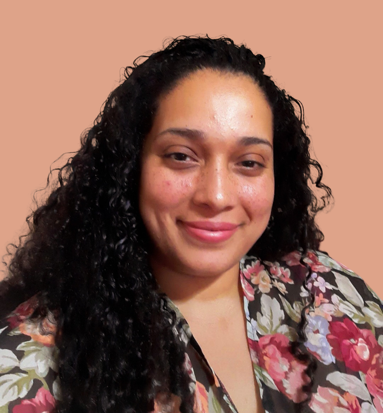 A woman with curly black hair smiling at the camera, wearing a floral shirt, in front of a plain beige background.