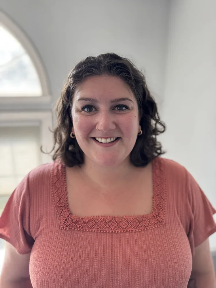 A woman with dark, curly hair smiling in front of a window with a white wall background. She is wearing a salmon-colored top with embroidered details around the neckline.