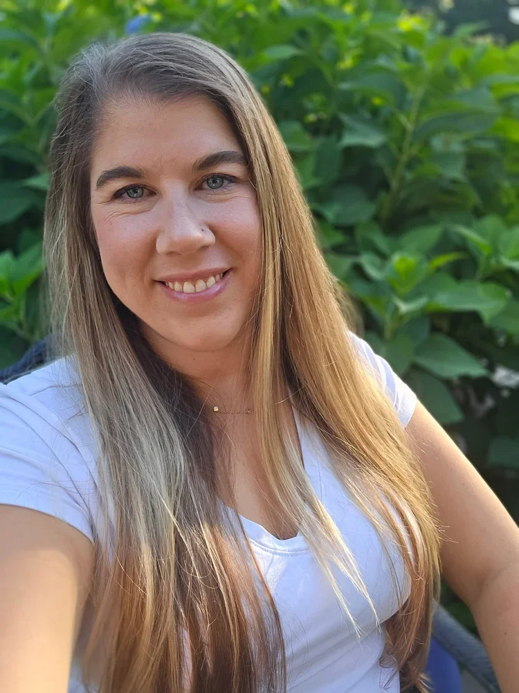 A woman with long blonde hair smiling outdoors in front of green leafy plants.