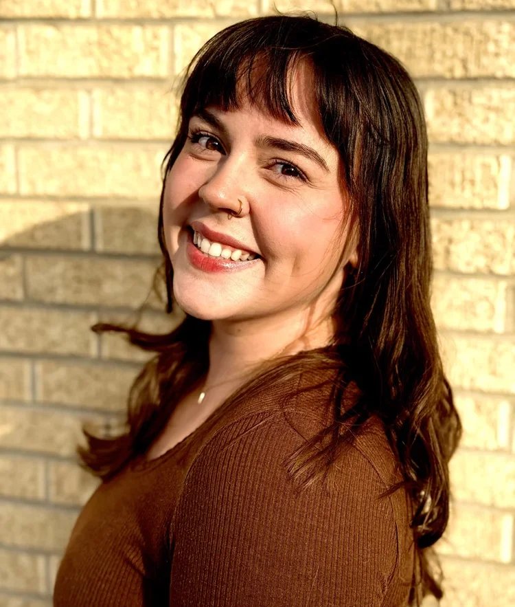 A woman with dark brown hair and a nose piercing smiling at the camera, standing against a brick wall.
