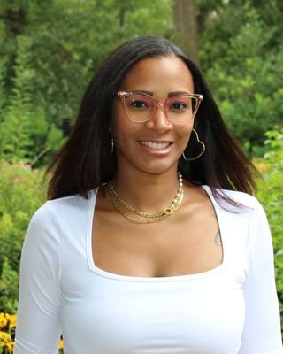 A woman with long dark hair, wearing glasses, a white top, gold jewelry, and hoop earrings, standing outdoors with green trees in the background.