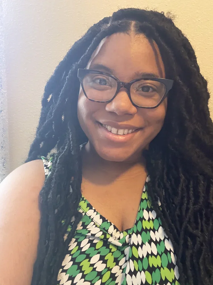 A young woman with long dreadlocks wearing glasses and a patterned sleeveless top, smiling at the camera.