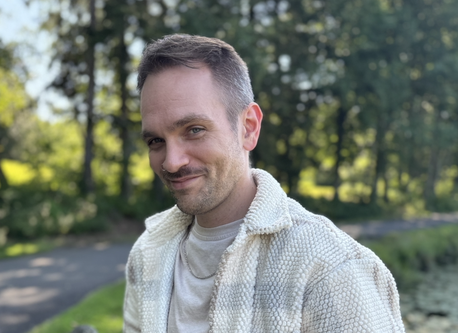 A man with short brown hair and a light beard, smiling outdoors with trees and a path in the background.