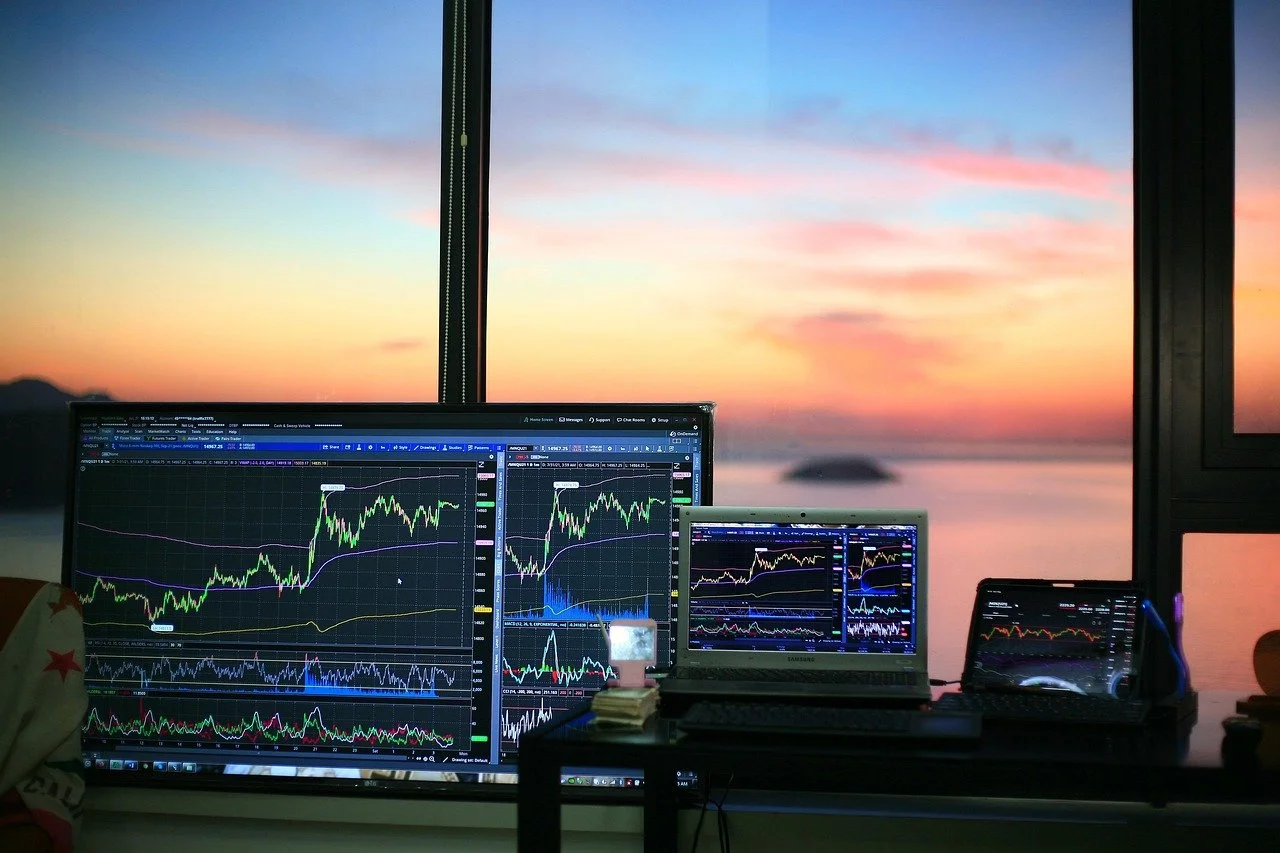 Multiple computer screens displaying stock market charts inside a room with a large window showing a sunset over the ocean.