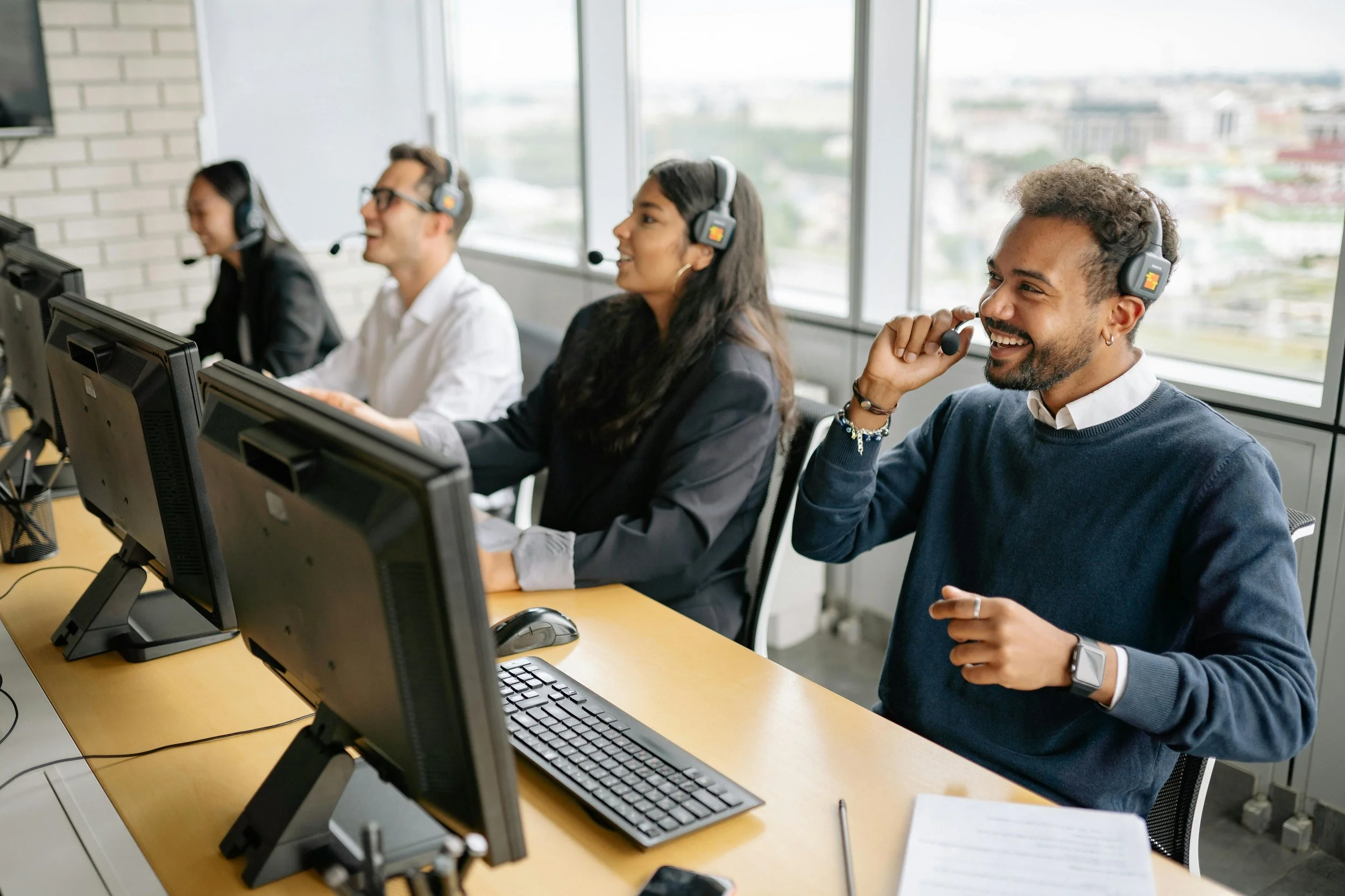 Four customer service representatives wearing headsets working at computers in an office with large windows showing a cityscape.