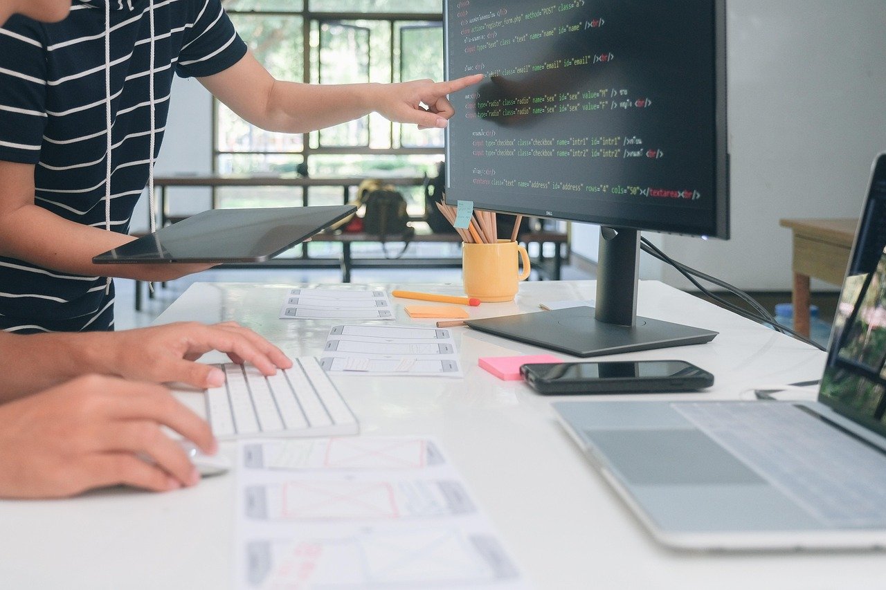 Two people working on web development, one pointing at a computer monitor displaying code and the other typing on a keyboard at a cluttered desk with various devices and papers.