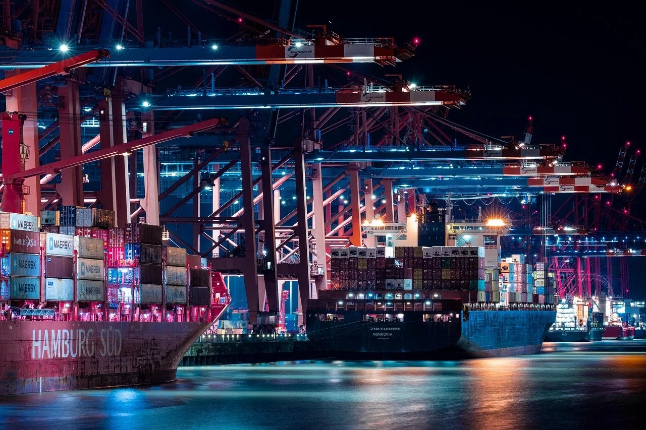 Night view of a busy container port with cranes and cargo ships docked and AI smart cameras, illuminated by bright lights.