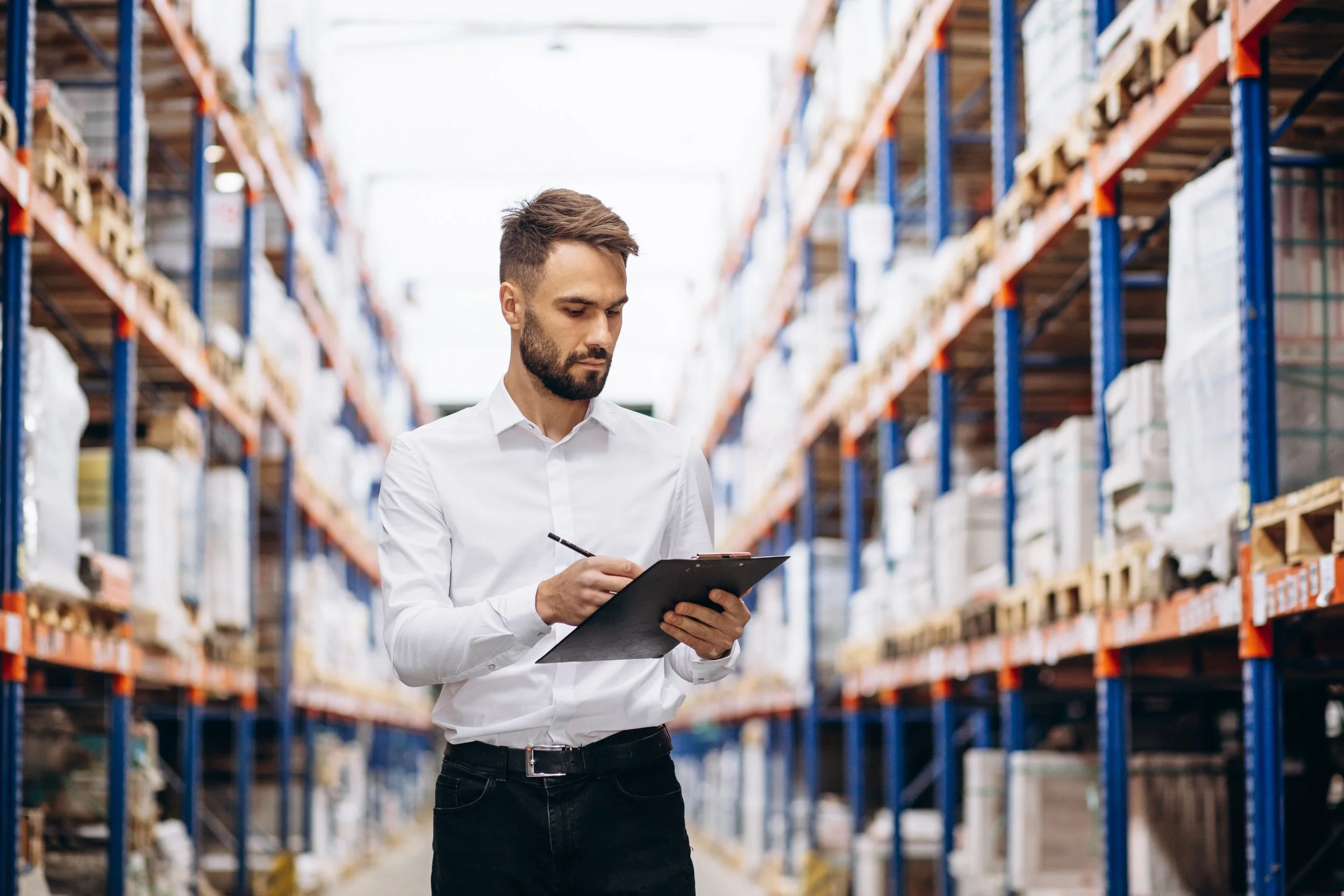 A local business man in a white shirt and black pants is standing in a warehouse aisle, holding a clipboard and writing.